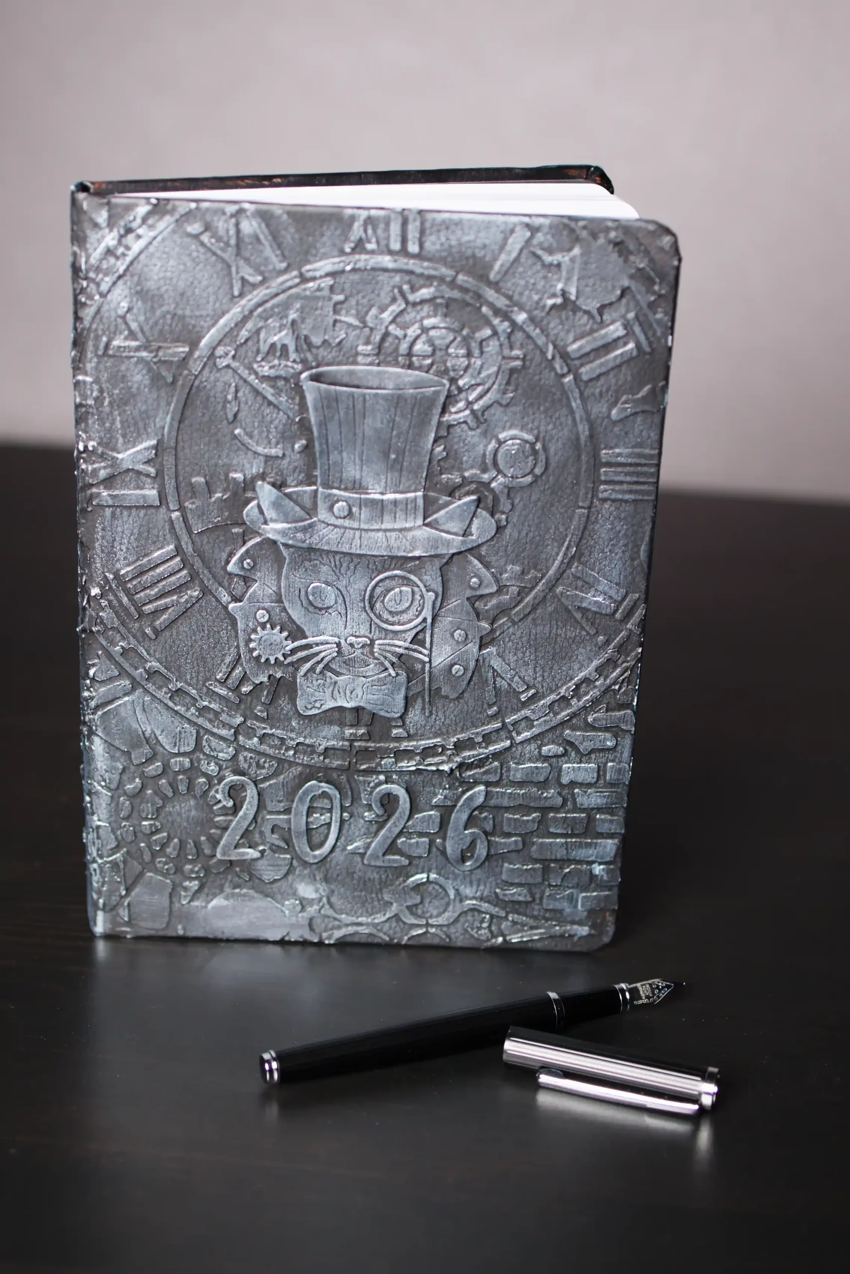 Agenda 2026 Steampunk : Couverture texturée gris métallique avec un chat stylisé portant un chapeau haut de forme et un monocle, sur fond d'horloge. Un stylo plume noir est posé devant l'agenda, sur une surface sombre.