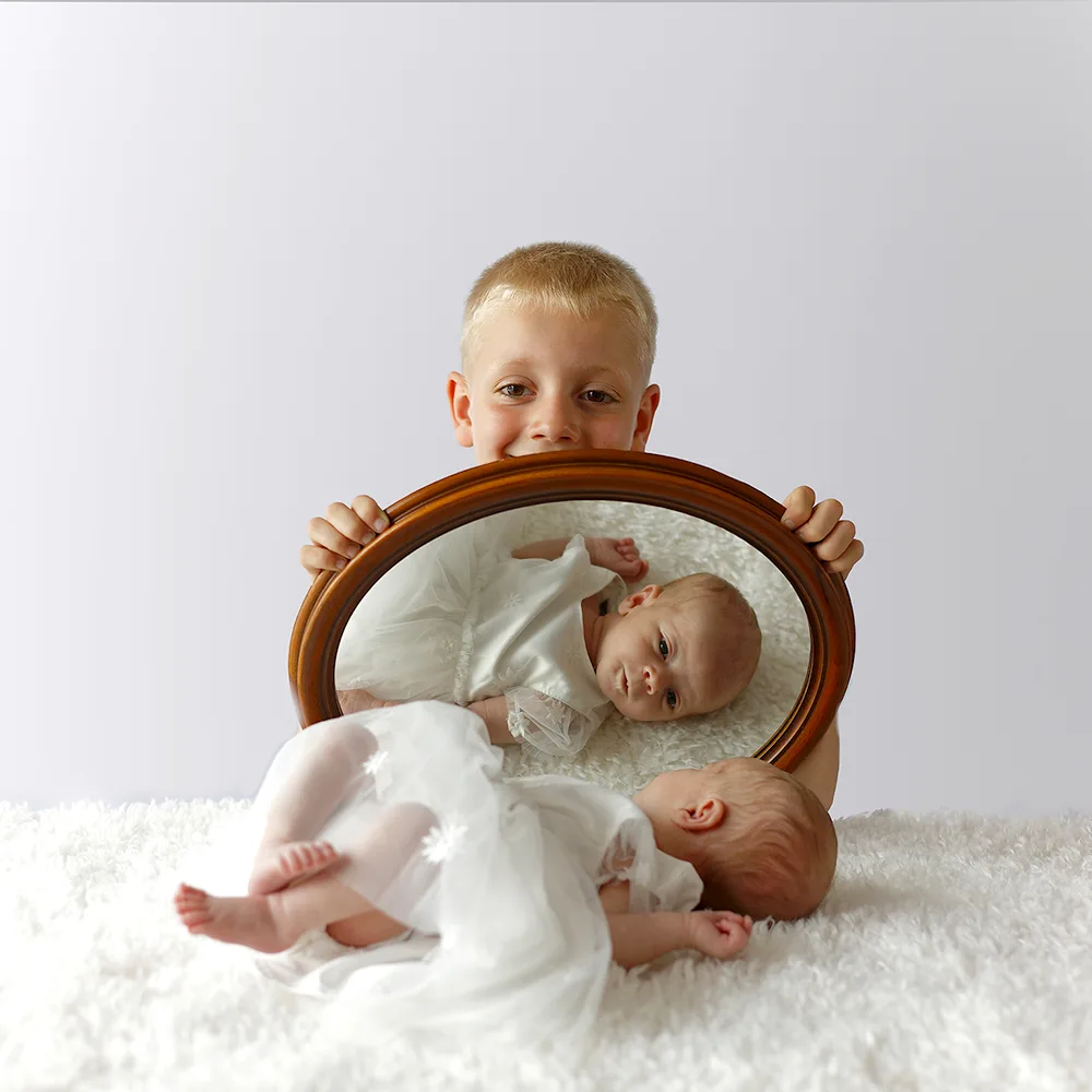  Un jeune garçon souriant tenant un miroir qui reflète sa petite sœur nouveau-née. Le bébé est allongé sur un tapis doux et blanc, vêtu d'une robe blanche assortie, créant une image tendre et familiale.