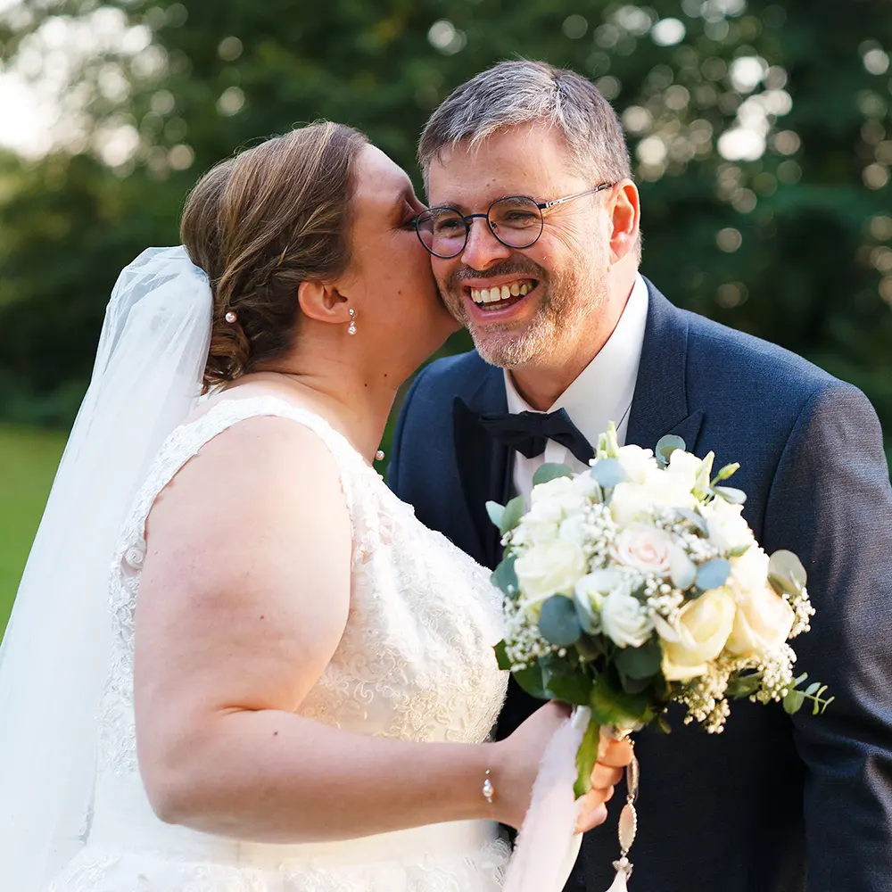 Moment de complicité des mariés | Photographe Mariage Arras Photographie de mariage d'un couple joyeux. La mariée murmure à l'oreille de son époux qui rit, un instant de bonheur capturé dans un parc verdoyant par un photographe de mariage près de Sainte-Catherine.