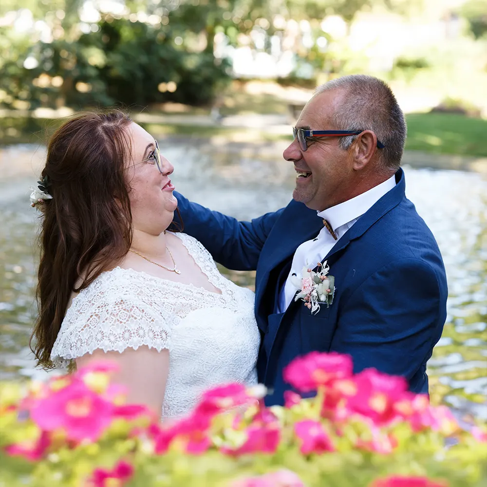 Photo de mariés souriants devant un bassin | Photographe Arras Photographie joyeuse d'un couple de mariés souriant et se regardant avec amour, devant un bassin dans un parc. Les fleurs roses au premier plan ajoutent une touche de couleur et de romantisme. Reportage photo de mariage dans le Pas-de-Calais à Hénin Beaumont