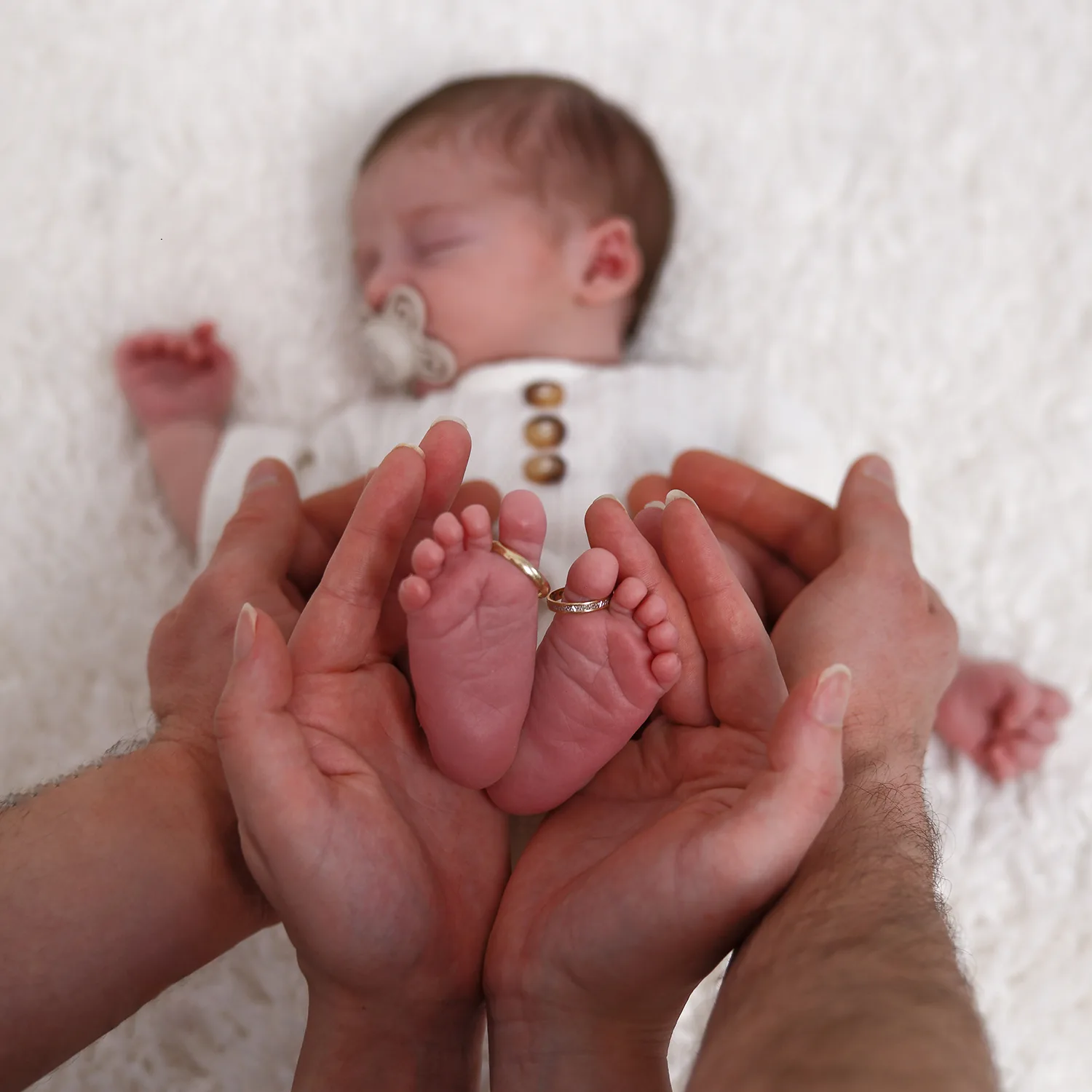 Les mains des parents tenant les pieds de leur bébé nouveau-né. Les alliances des parents sont passées autour des orteils du bébé, symbolisant le lien d'amour familial. Le bébé dort paisiblement en arrière-plan, rendant l'image tendre et personnelle.