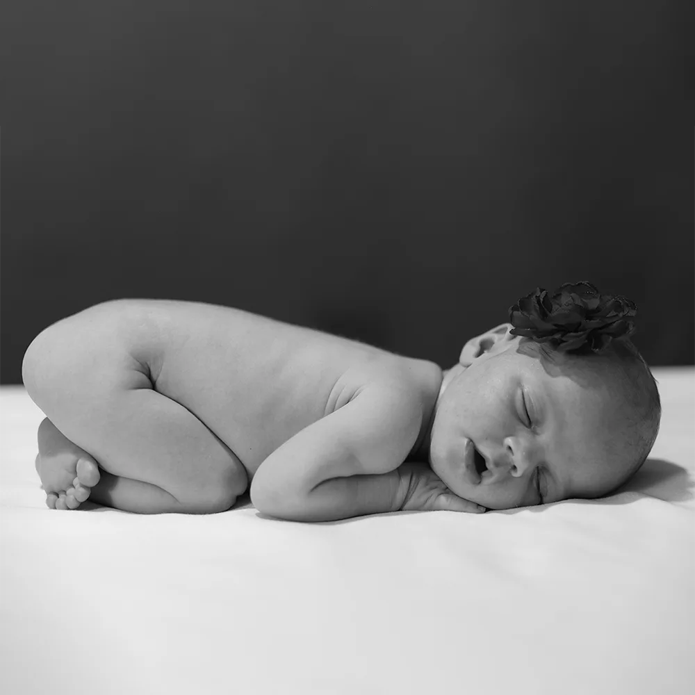 Photo en noir et blanc d'un nouveau-né endormi sur le ventre. Le bébé porte une petite fleur foncée sur l'oreille. L'image met en valeur la douceur et la fragilité des premiers jours de vie.