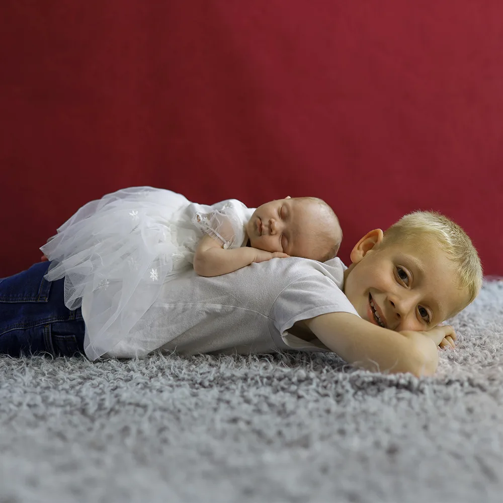 Un grand frère souriant, allongé sur le ventre, avec sa petite sœur nouveau-née endormie sur son dos. Le garçon regarde l'objectif, tandis que le bébé repose en toute sécurité, illustrant le lien fort entre frères et sœurs.