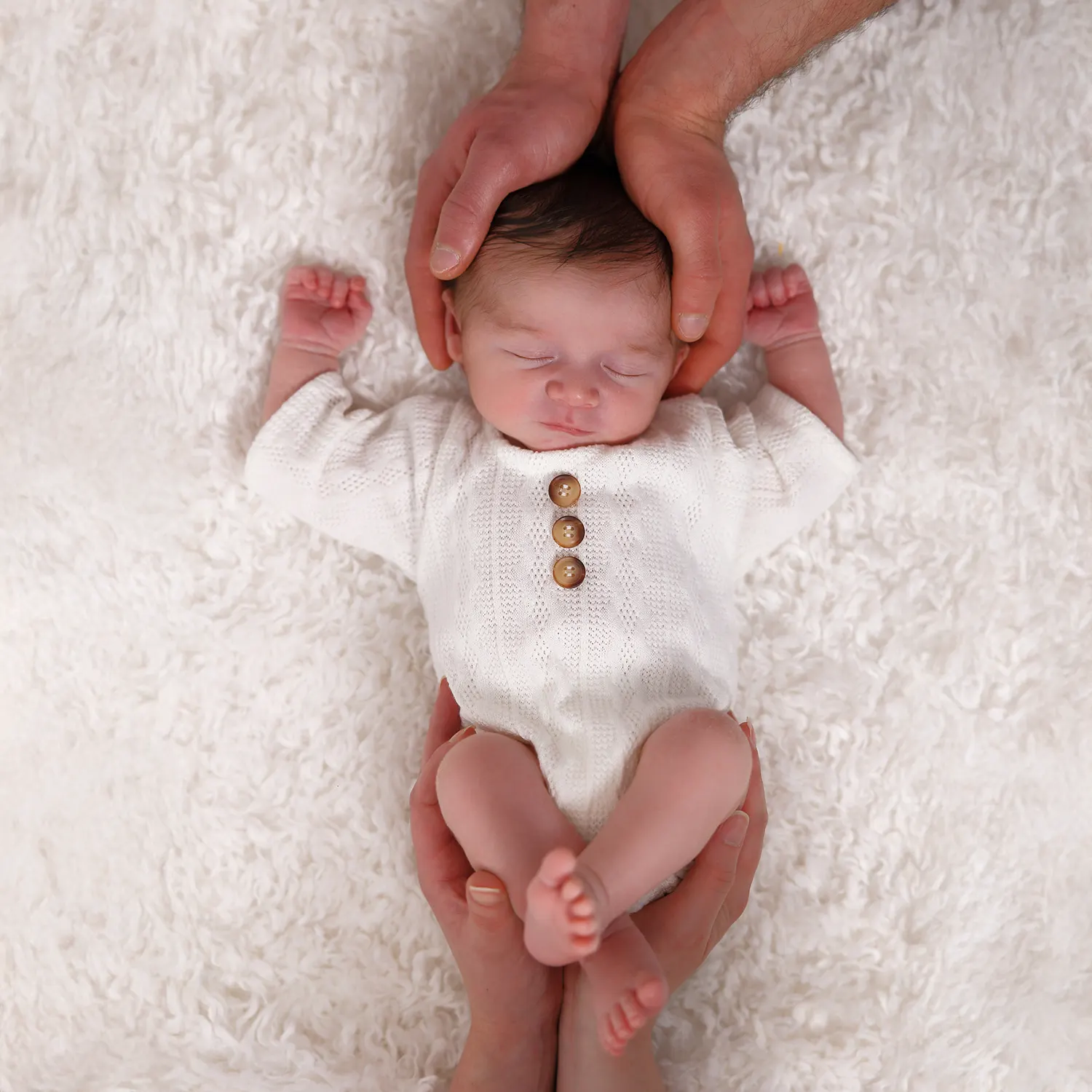 Vue de dessus d'un nouveau-né endormi, habillé d'une tenue en tricot blanche. Les mains des parents encadrent délicatement sa tête et ses pieds, symbolisant la protection et l'amour familial sur un fond blanc et doux.