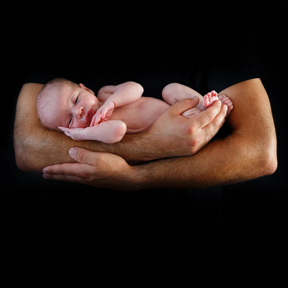 Les bras d'un parent tenant délicatement un nouveau-né sur un fond noir. Le bébé est nu, reposant en toute sécurité, soulignant la fragilité et la protection offertes par les parents.
