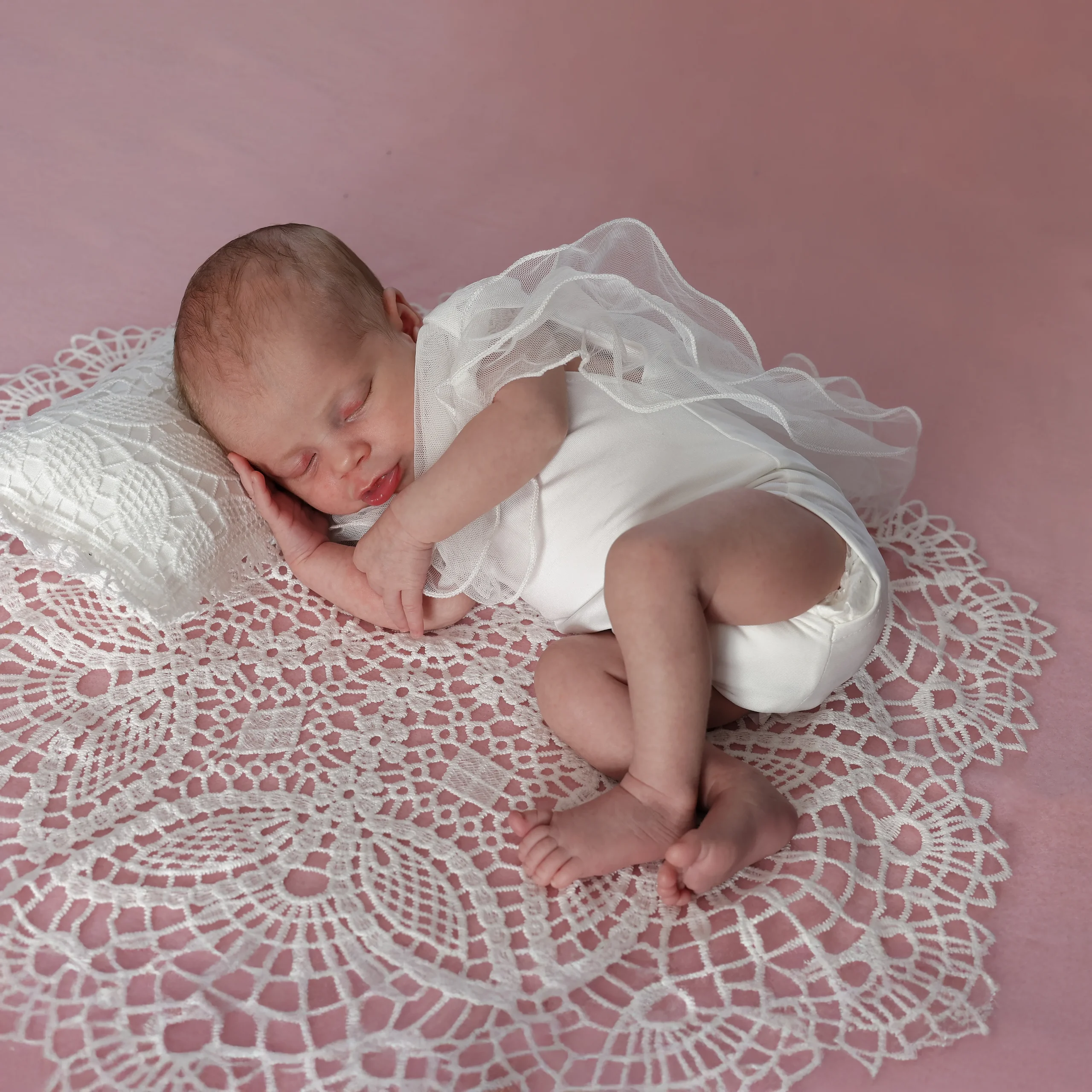 bébé allongé sur le coté. Elle est allongée sur un napperon rond brodé. L'oreiller est recouvert de broderie. Elle porte un body avec du voile.