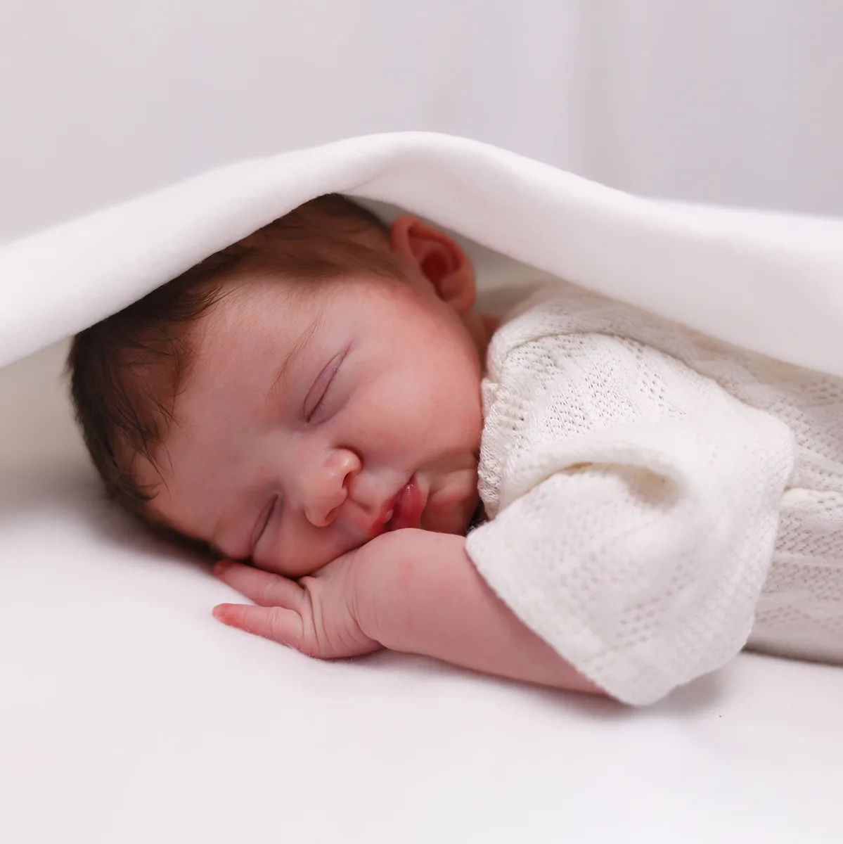 Gros plan d'un nouveau-né paisiblement endormi, la tête et le corps partiellement recouverts d'un drap blanc. Le bébé a l'air serein, sa petite main sous le menton, illustrant la tranquillité d'un sommeil de nouveau-né.