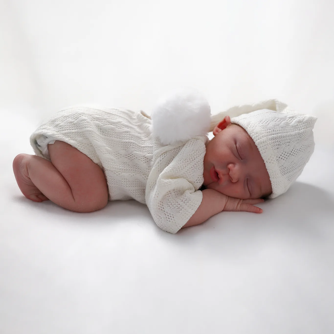 Un nouveau-né endormi sur le côté, vêtu d'une jolie combinaison en tricot et d'un bonnet assorti, de couleur blanche. L'enfant dort paisiblement sur un fond blanc, mettant en valeur la douceur de la tenue.
