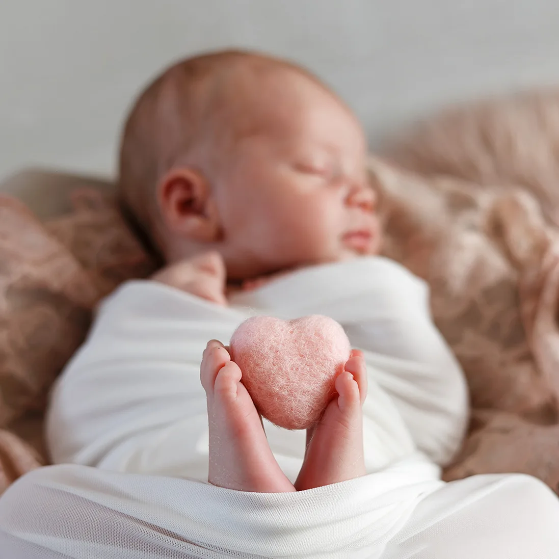 Les petits pieds d'un nouveau-né enveloppé dans un tissu blanc, tenant délicatement un petit cœur rose en feutre. Le bébé dort paisiblement en arrière-plan, créant une image de douceur et d'amour.
