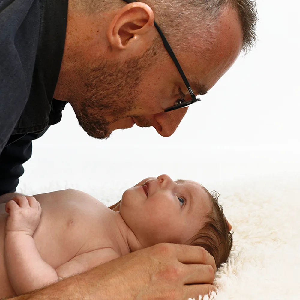 Gros plan sur un père penché sur son bébé nouveau-né. Le père regarde l'enfant avec tendresse, tandis que le bébé regarde son père, les deux visages se faisant face. L'image capture le lien affectif entre le père et son enfant sur un fond doux.