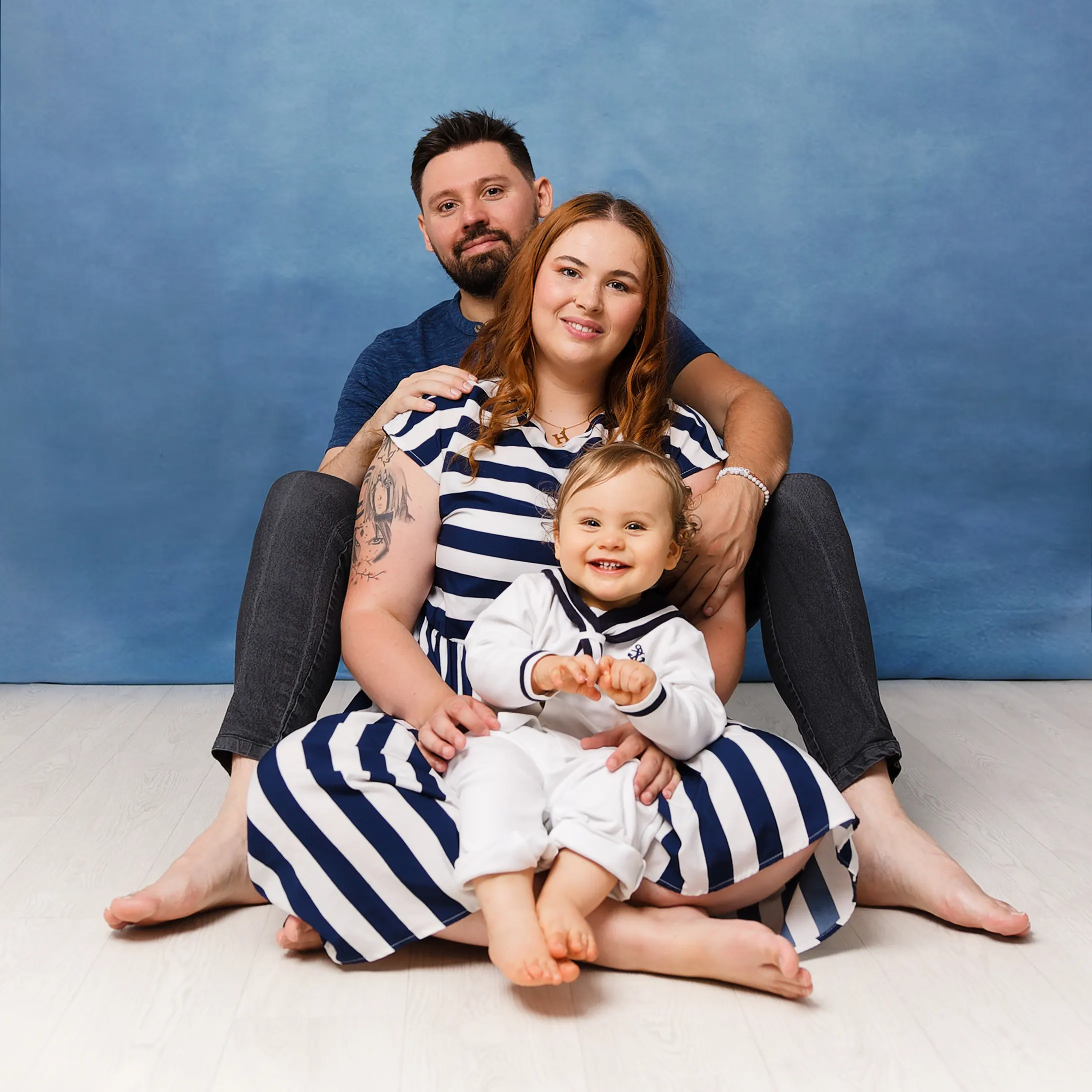 Famille de trois personnes assise sur le sol en studio, le bébé en tenue de marin est assis devant sa mère, tous regardent la caméra en souriant. Photo de famille à Sainte-Catherine.