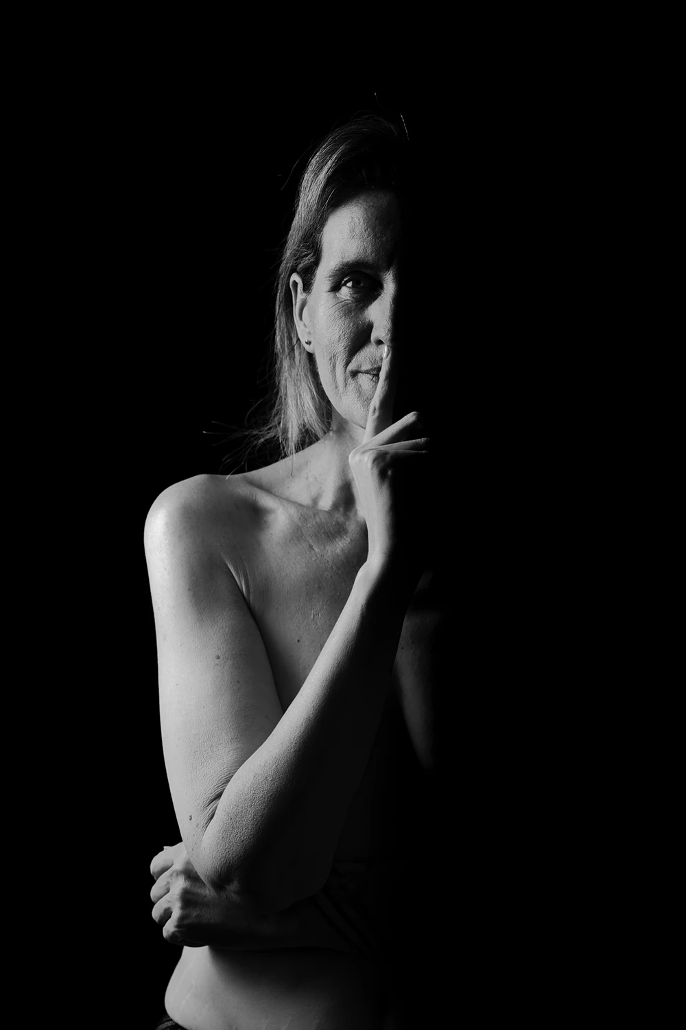 Portrait artistique boudoir en noir et blanc d'une femme mature avec le doigt sur les lèvres, évoquant le secret et l'intimité. Éclairage dramatique mettant en valeur le visage et l'épaule. Séance réalisée par votre photographe à Sainte-Catherine, Hauts-de-France.