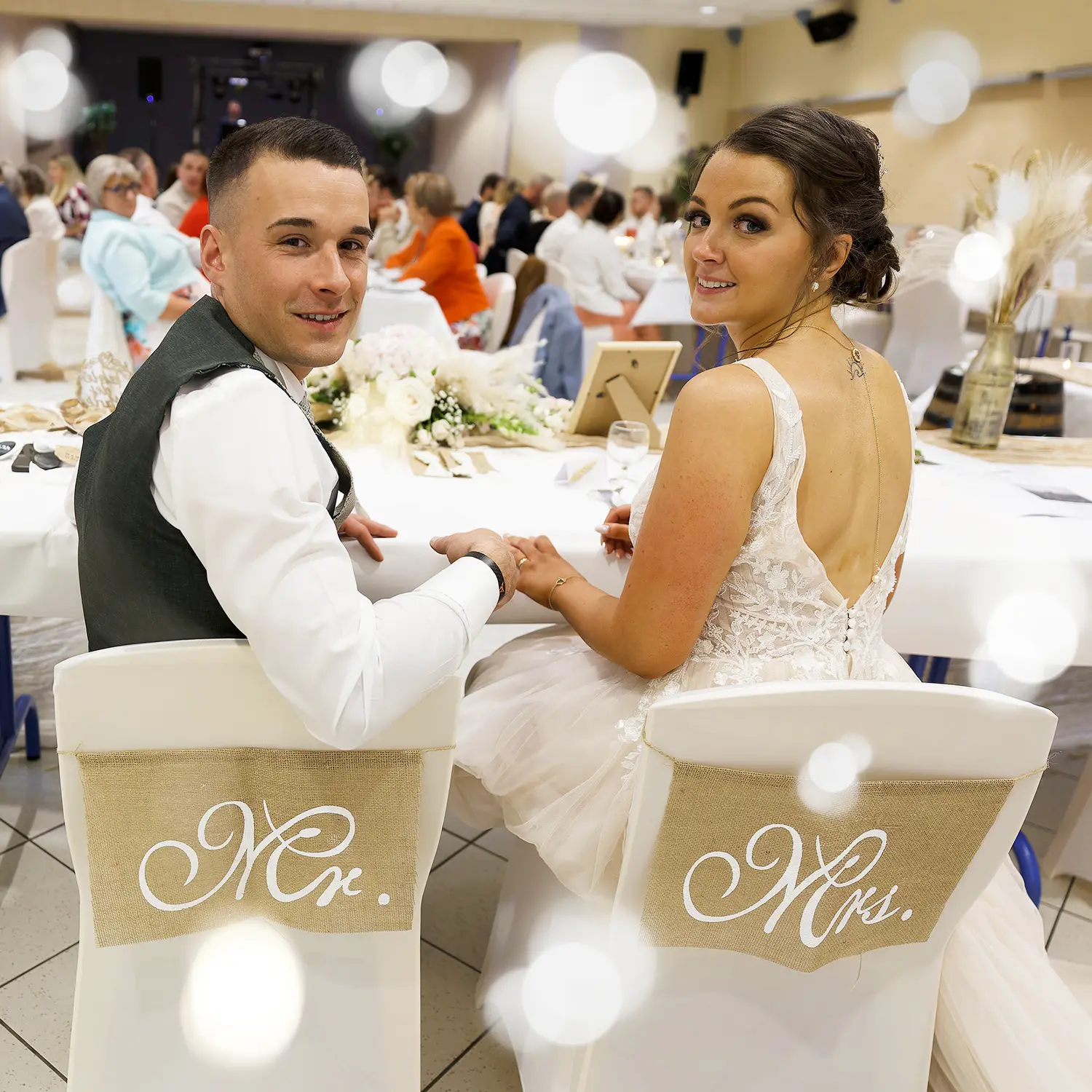 Photos de soirée de mariage | Photographe Arras Manon et Vincent, les jeunes mariés, posent à leur table de réception lors de la soirée de leur mariage. Les chaises Mr. et Mrs. sont décorées pour l'occasion. Photographe de mariage dans la région de Lens, dans la salle des fêtes à Souchez