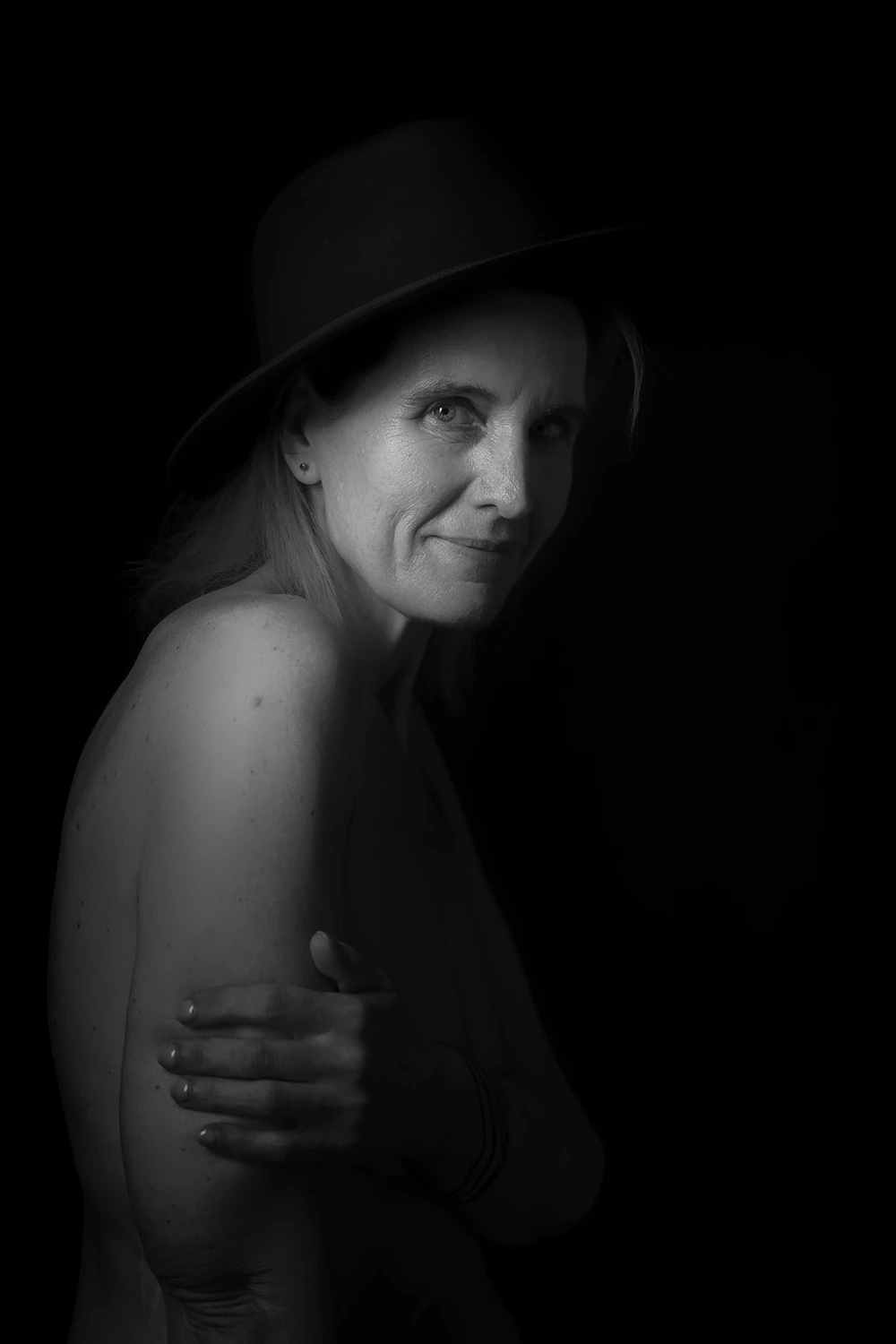 Photographie boudoir en noir et blanc d'une femme mature posant avec un chapeau noir, regardant par-dessus son épaule. L'éclairage met en valeur son expression douce et sa peau. Séance réalisée par votre photographe à Sainte-Catherine, près d'Arras.