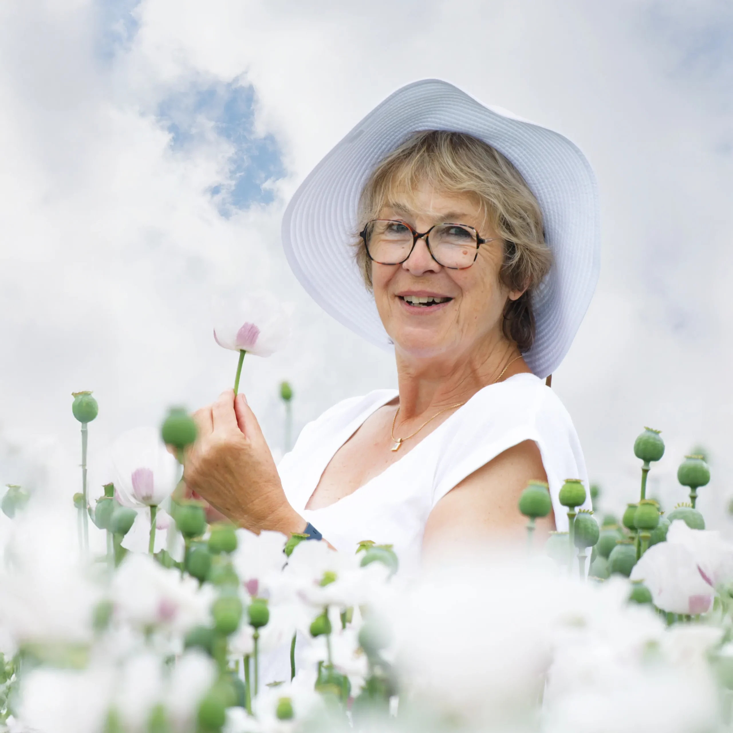 Portrait de femme souriante tenant un pavot blanc, photo en extérieur au milieu d'un champ de fleurs à Sainte-Catherine.