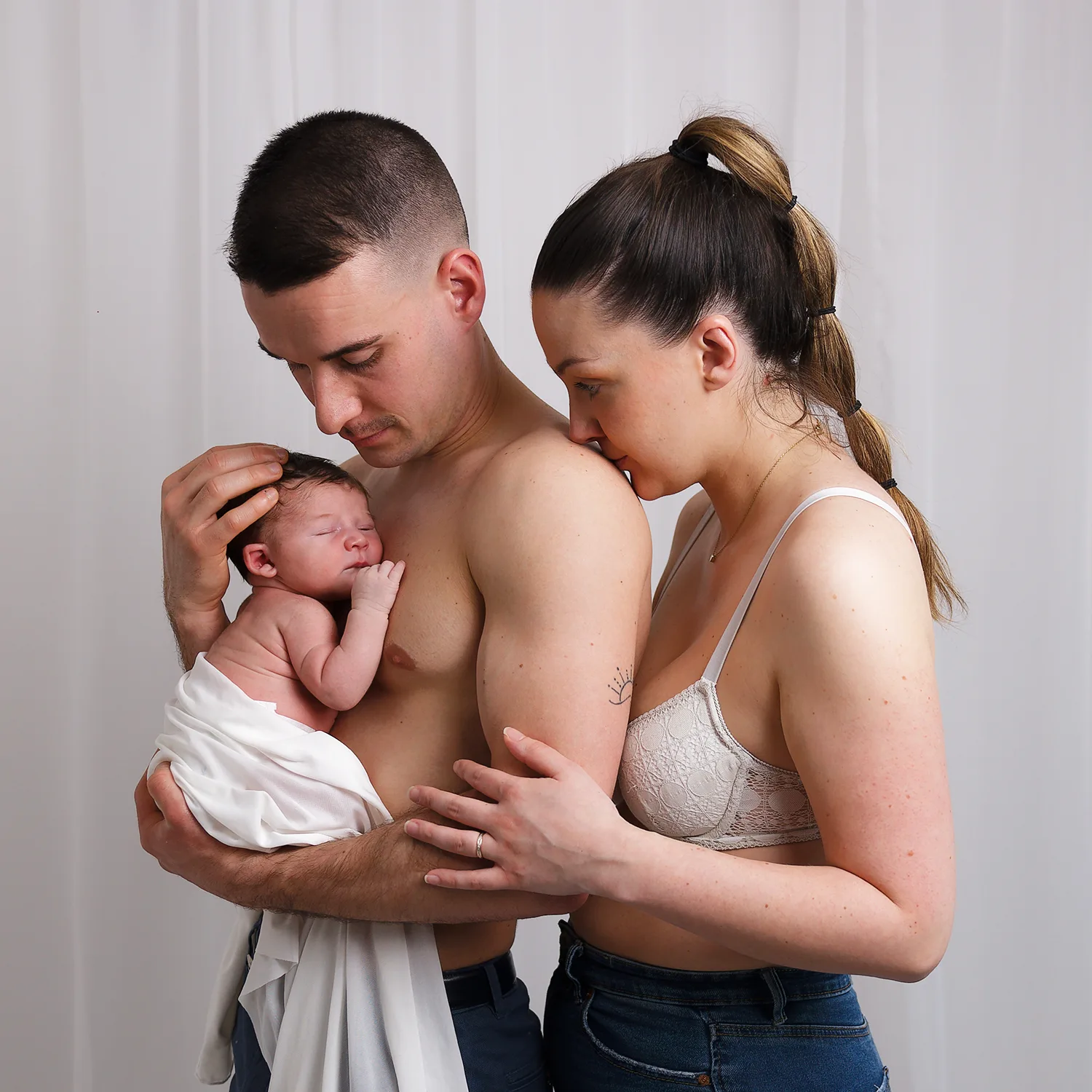 Les parents tiennent leur nouveau-né dans leurs bras, montrant un moment de tendresse familiale. Le père berce le bébé, tandis que la mère pose sa tête sur son épaule. L'image capture le lien affectueux et protecteur de cette jeune famille.