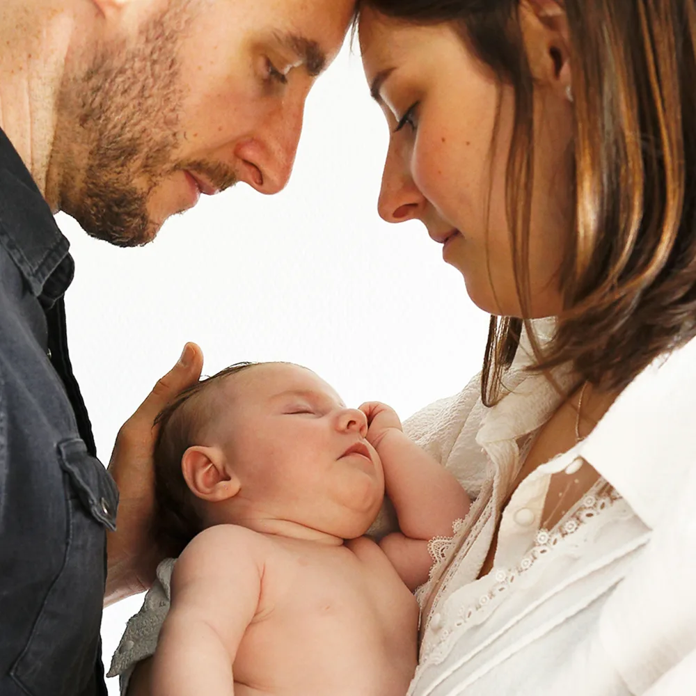 Les parents se penchent l'un vers l'autre, les fronts se touchant, en tenant leur nouveau-né endormi. L'image est un portrait intime qui illustre le lien fort et l'amour partagé par cette nouvelle famille.