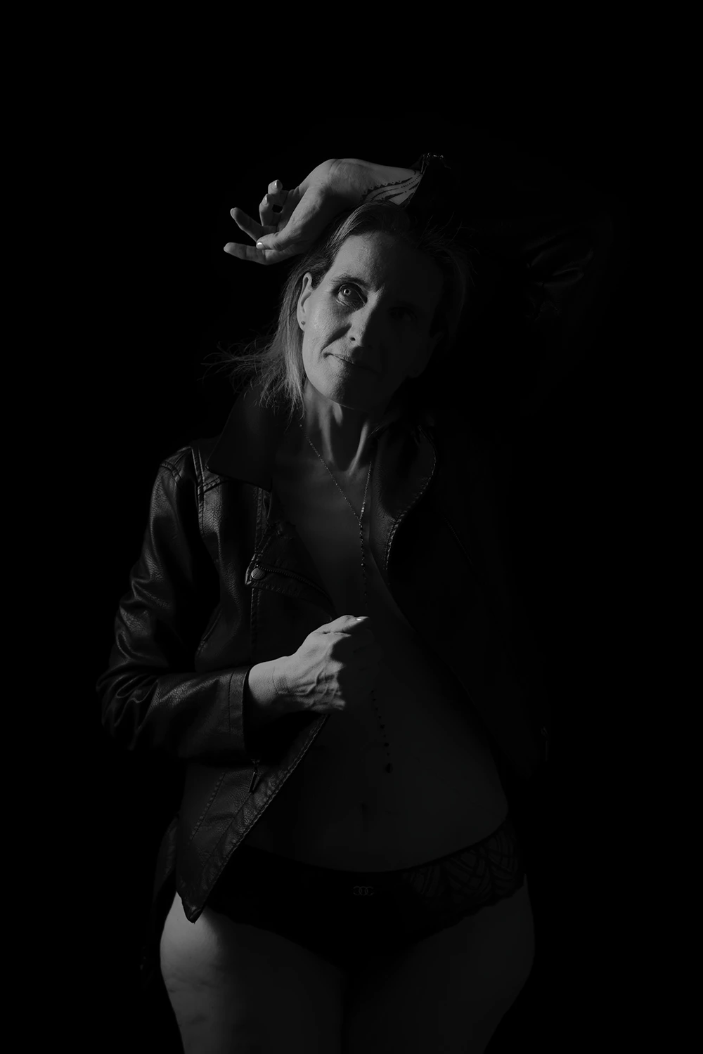 Photo de boudoir en noir et blanc d'une femme mature posant en veste en cuir ouverte et culotte, les bras levés, transmettant force et confiance. Mise en lumière du ventre et des détails de la veste. Réalisée par votre photographe spécialisée à Sainte-Catherine, près d'Arras