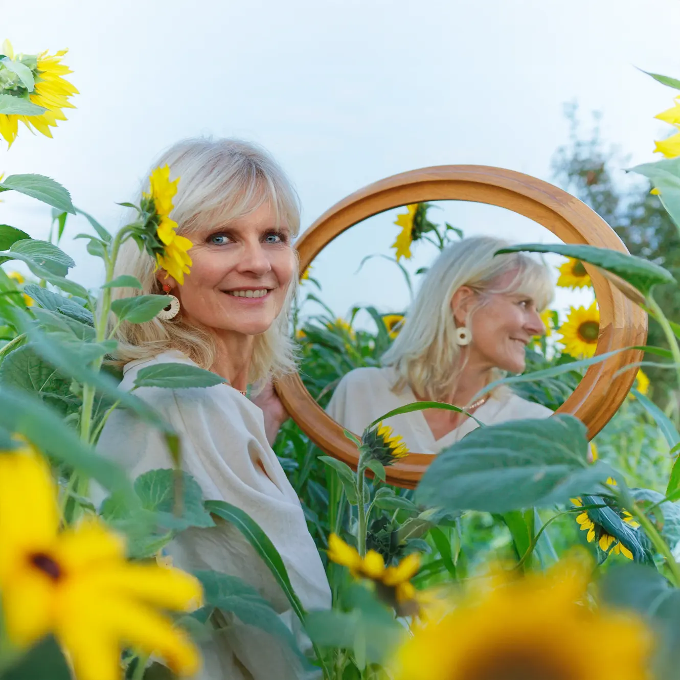 Portrait créatif d'une femme souriante dans un champ de tournesols, tenant un miroir rond, séance photo à Sainte-Catherine.