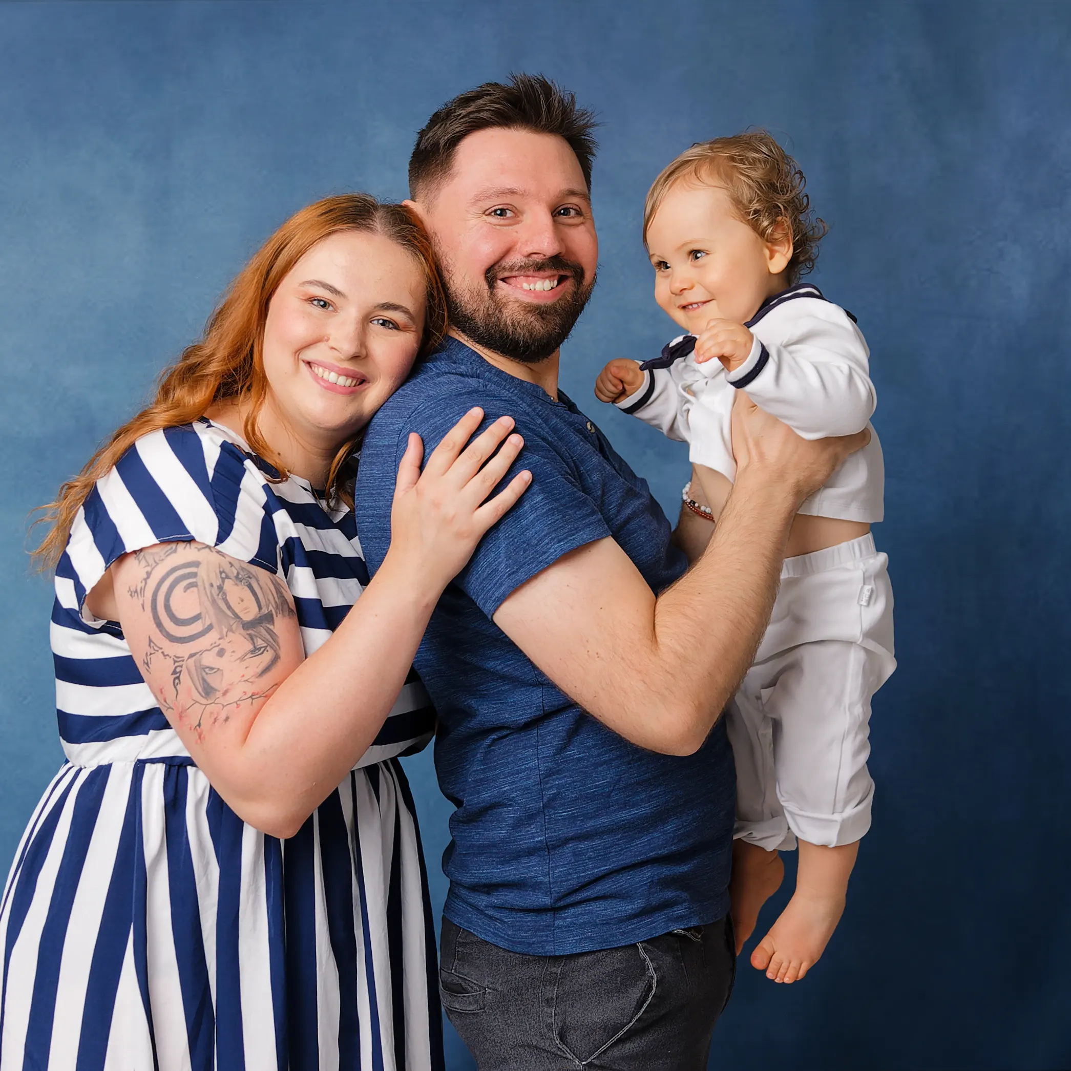 Portrait de famille en studio, le père porte le bébé en tenue de marin, la mère l'enlace affectueusement, tous trois souriants. Photo par un photographe à Sainte-Catherine