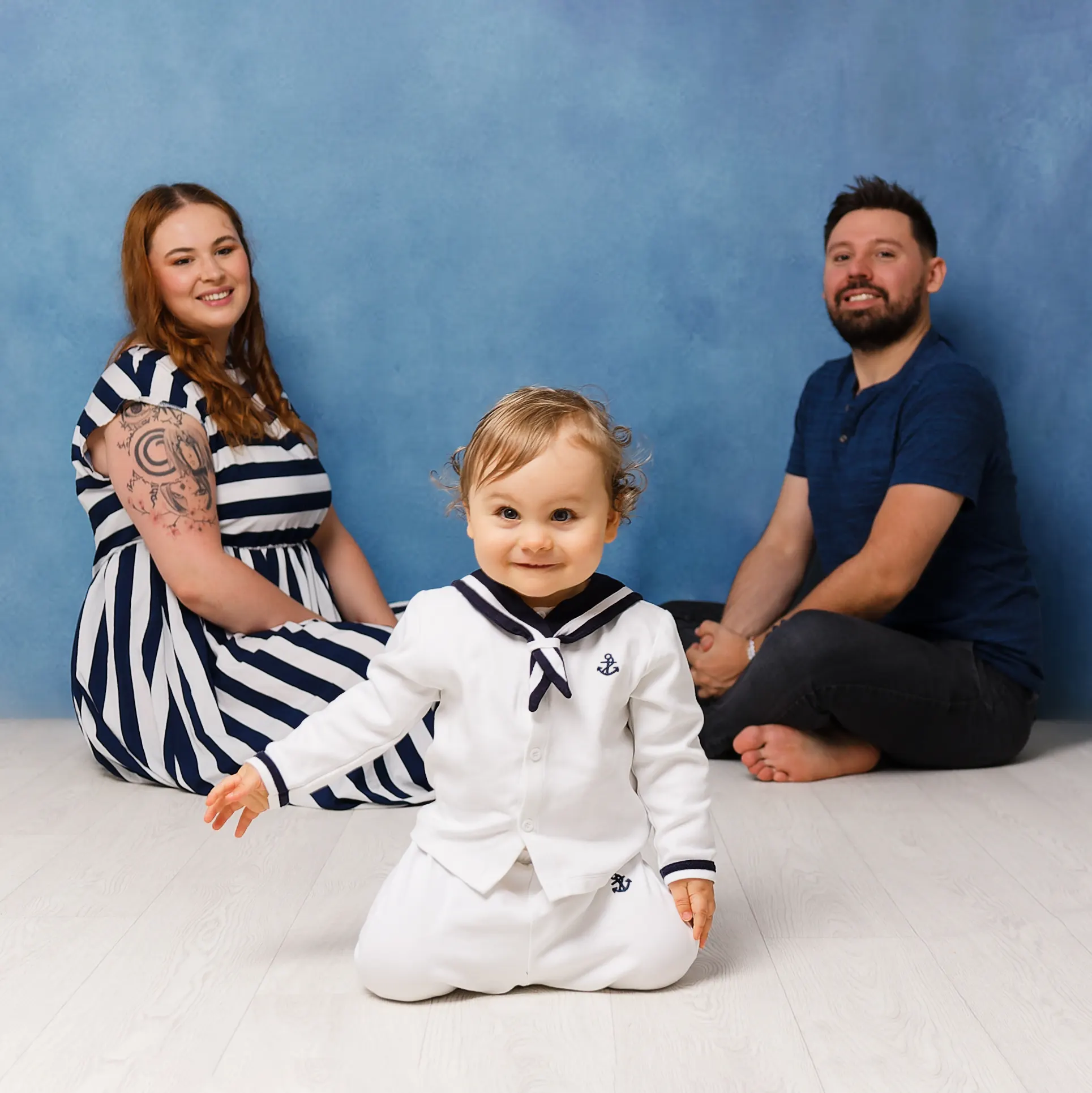 Photo de famille joyeuse avec un bébé en tenue de marin au premier plan, souriant à la caméra, et ses parents derrière, prise par un photographe de portraits à Sainte-Catherine