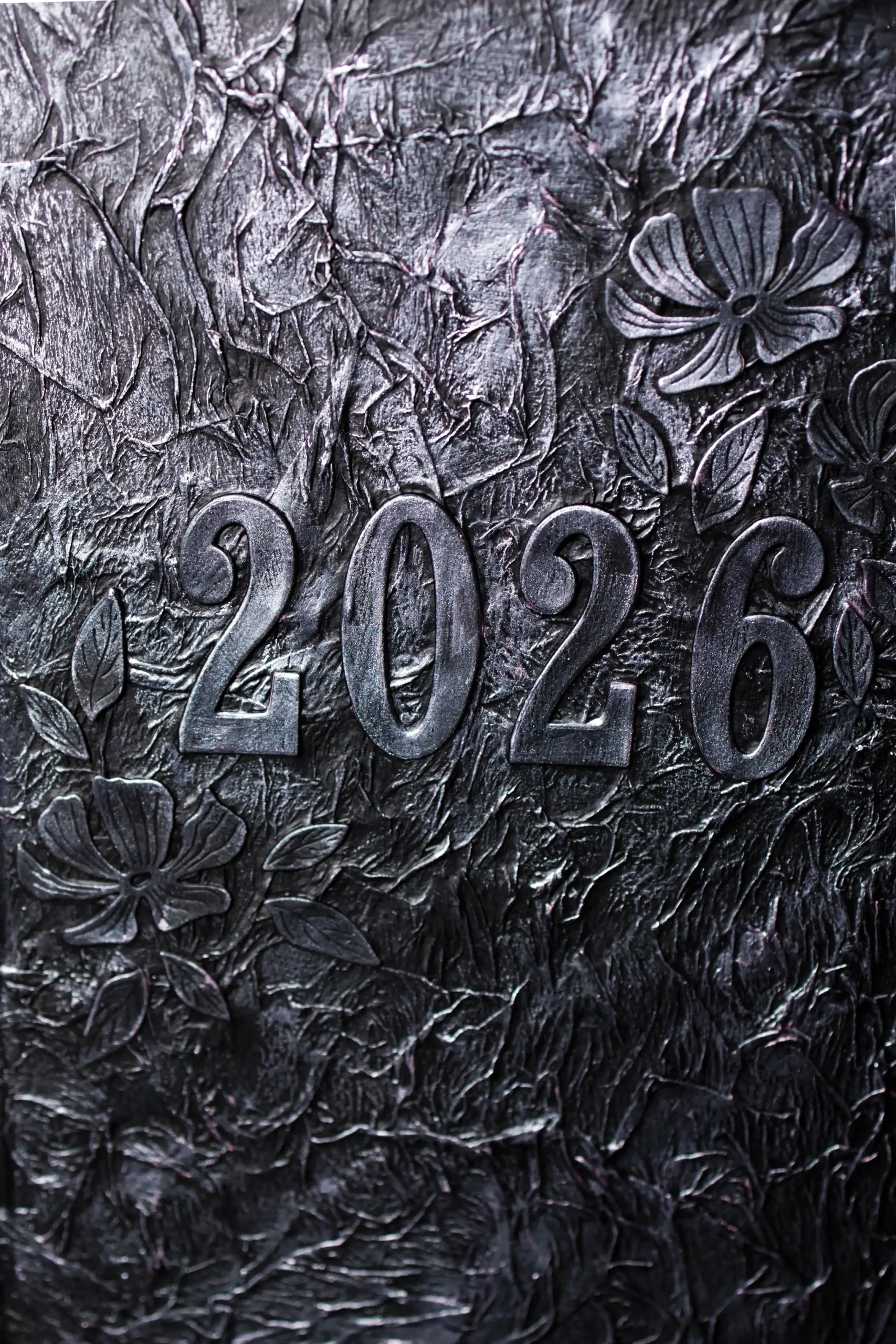 Gros plan sur le nombre 2026 de l'agenda, dans un aspect métallique argenté, incrusté sur un fond texturé et sombre, évoquant une matière travaillée ou une écorce. Des motifs floraux sculptés sont visibles autour des chiffres.