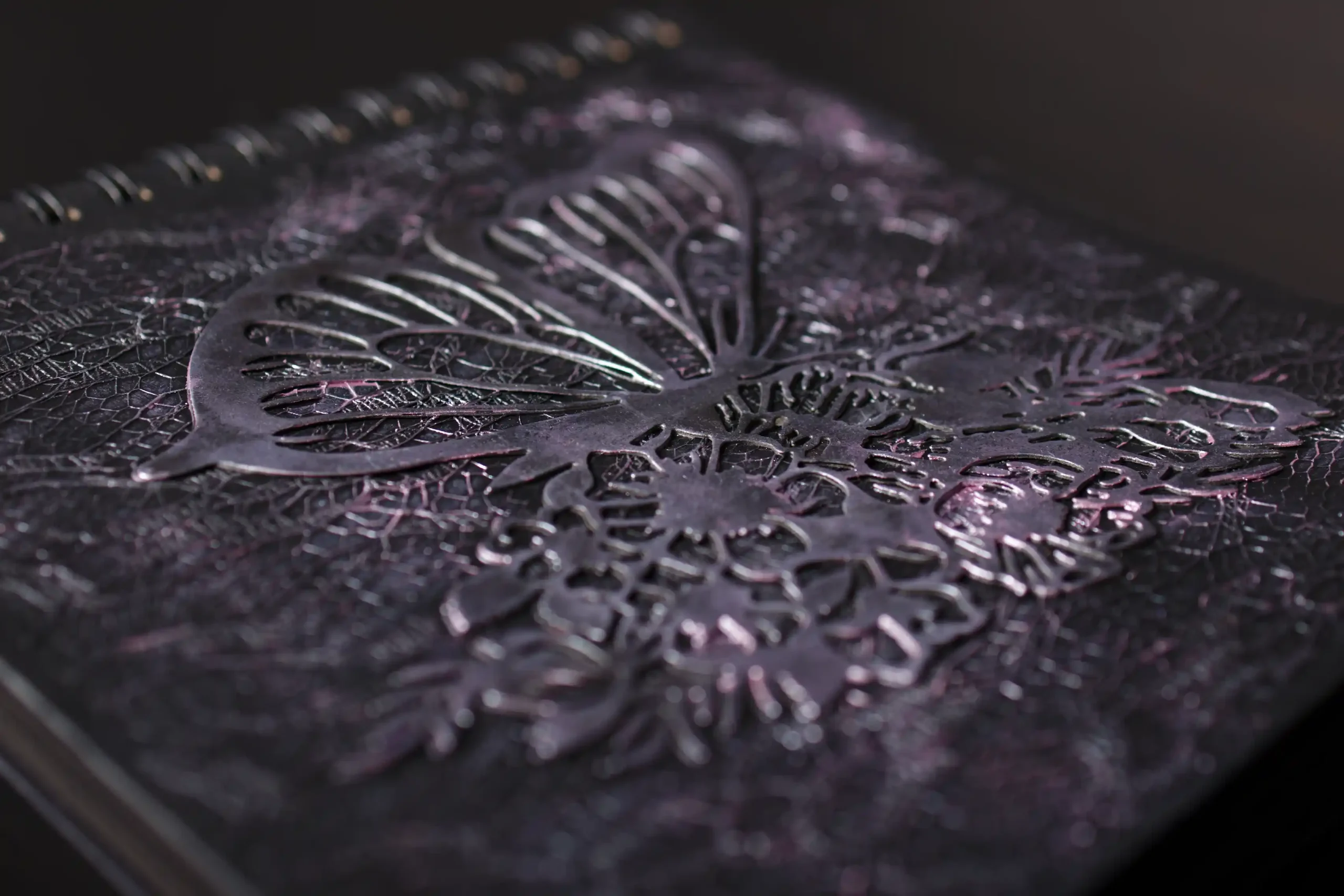 Gros plan en macro sur la couverture d'un album photo noir et texturé. Un motif de papillon et de fleurs découpé en métal foncé est posé en relief, soulignant la texture craquelée de la couverture.