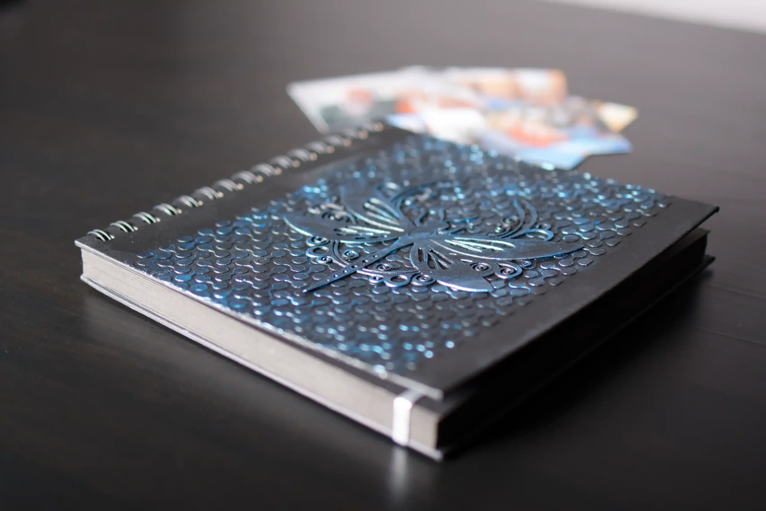 Gros plan sur un album photo ou livre d'or à spirales, posé sur une surface sombre. La couverture est noir mat avec un motif de libellule et des vagues en relief bleu métallique brillant. Quelques photos floues sont visibles en arrière-plan.