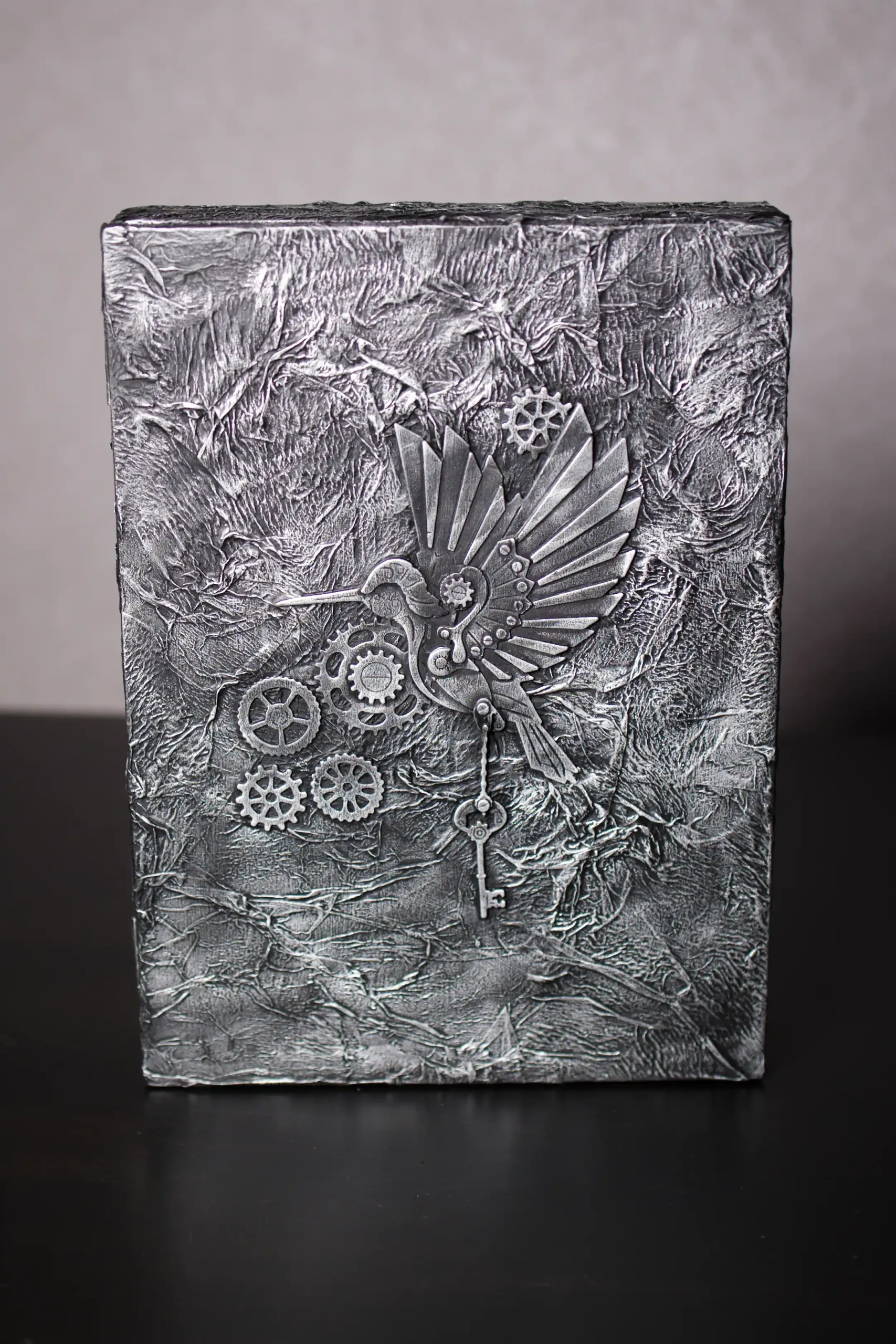 Boîte à clés de style Steampunk, à la texture très travaillée, gris et argenté. Un colibri mécanique avec des ailes en engrenages est fixé au centre, tenant une clé ancienne.