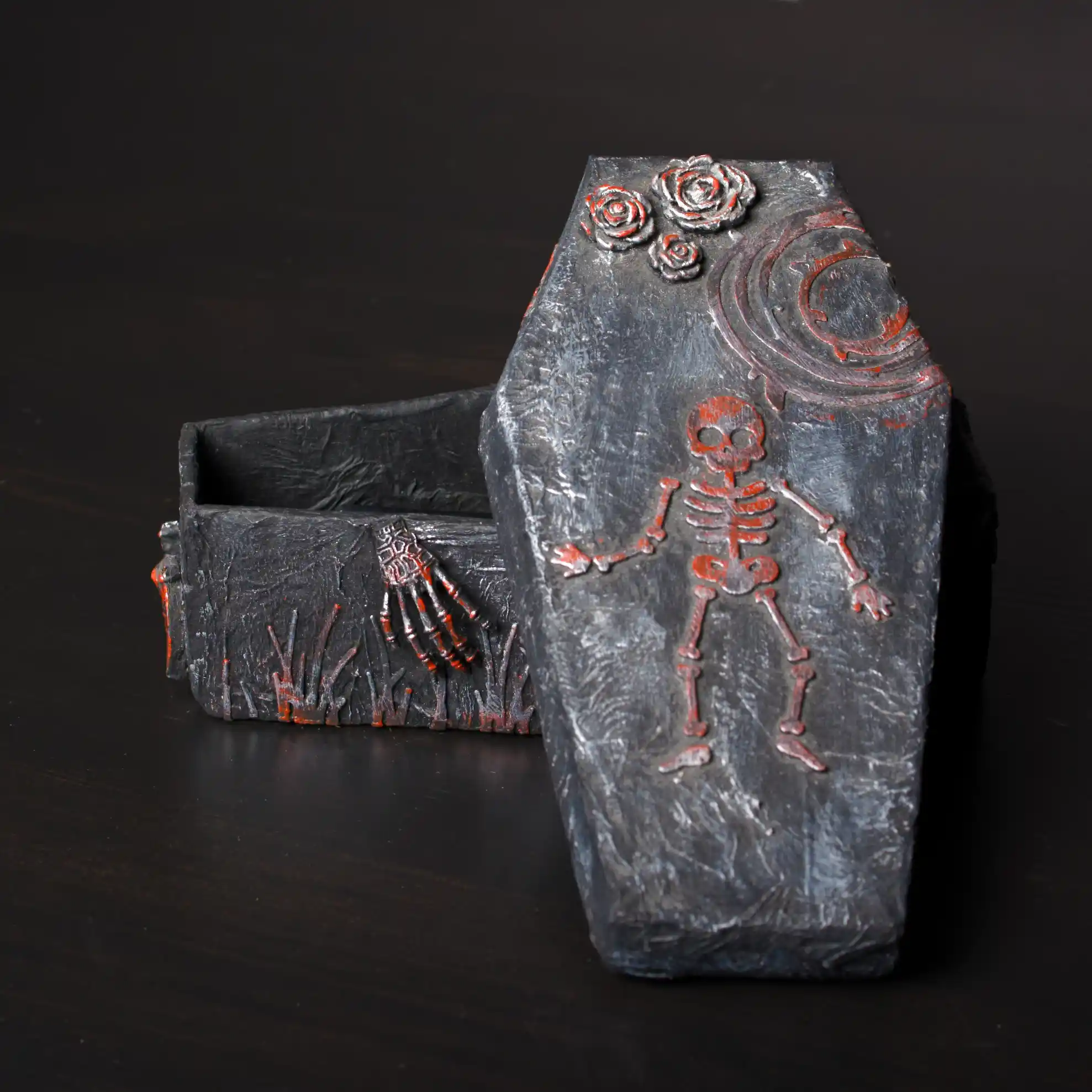 Boîte décorative en forme de cercueil, peinte en gris texturé et noir, ouverte sur le dessus. Le couvercle est orné d'un squelette en relief et de roses rouges et d'une lune peints. Le style est gothique.