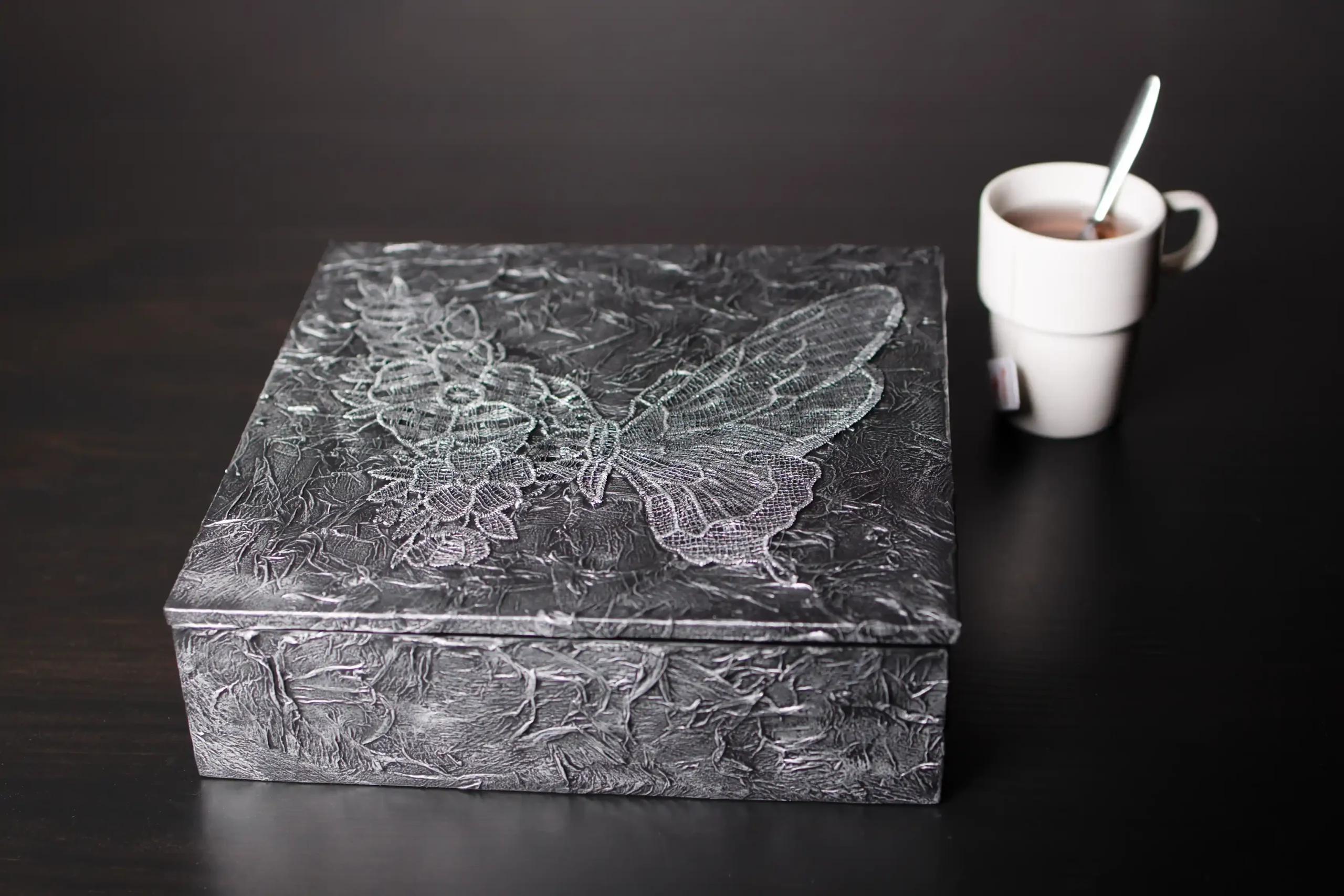 Boîte rectangulaire noire texturée, vraisemblablement pour le thé, avec un grand motif de papillon et de fleurs en relief métallique argenté. Une tasse de thé blanche avec un sachet est visible sur la droite, sur un fond sombre.