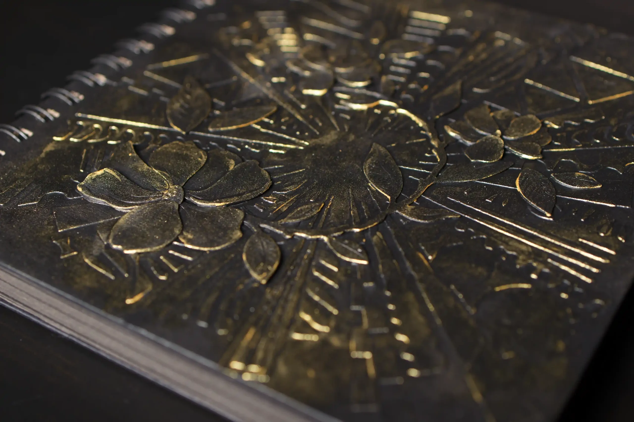 Gros plan sur la couverture d'un album photo à spirales noir. Le motif floral est en relief, avec des détails dorés brillants sur les fleurs et les feuilles, contrastant avec la texture sombre de la couverture.