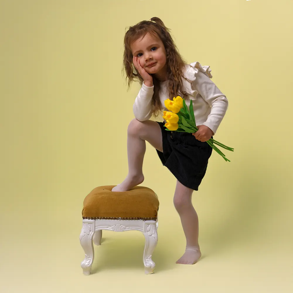 Portrait enfant de 3 ans posant dans un studio sur fond jaune. Elle a un pied appuyé sur un tbouret. Elle tient un bouquet de jonquilles. Cette photo a un esprit printanier.