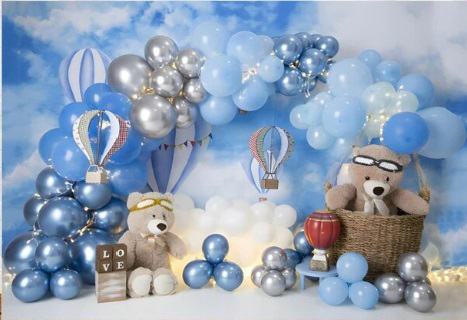 Fond photo représentant une scène pour un anniversaire d'enfant. Le décor est bleu, avec des ballons bleus er argentés. Il y a des nounours, dont 1 est dans un panier d'osier. Des petites montgolfières se trouvent aussi dans le décor.