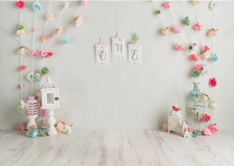 Fond photo pour un anniversaire d'un enfant. Le mur et le sol sont blancs. Des fleurs de papiers sont disposées en guirlandes. Des lampes sont posées sur le sol. les couleurs sont tendres.