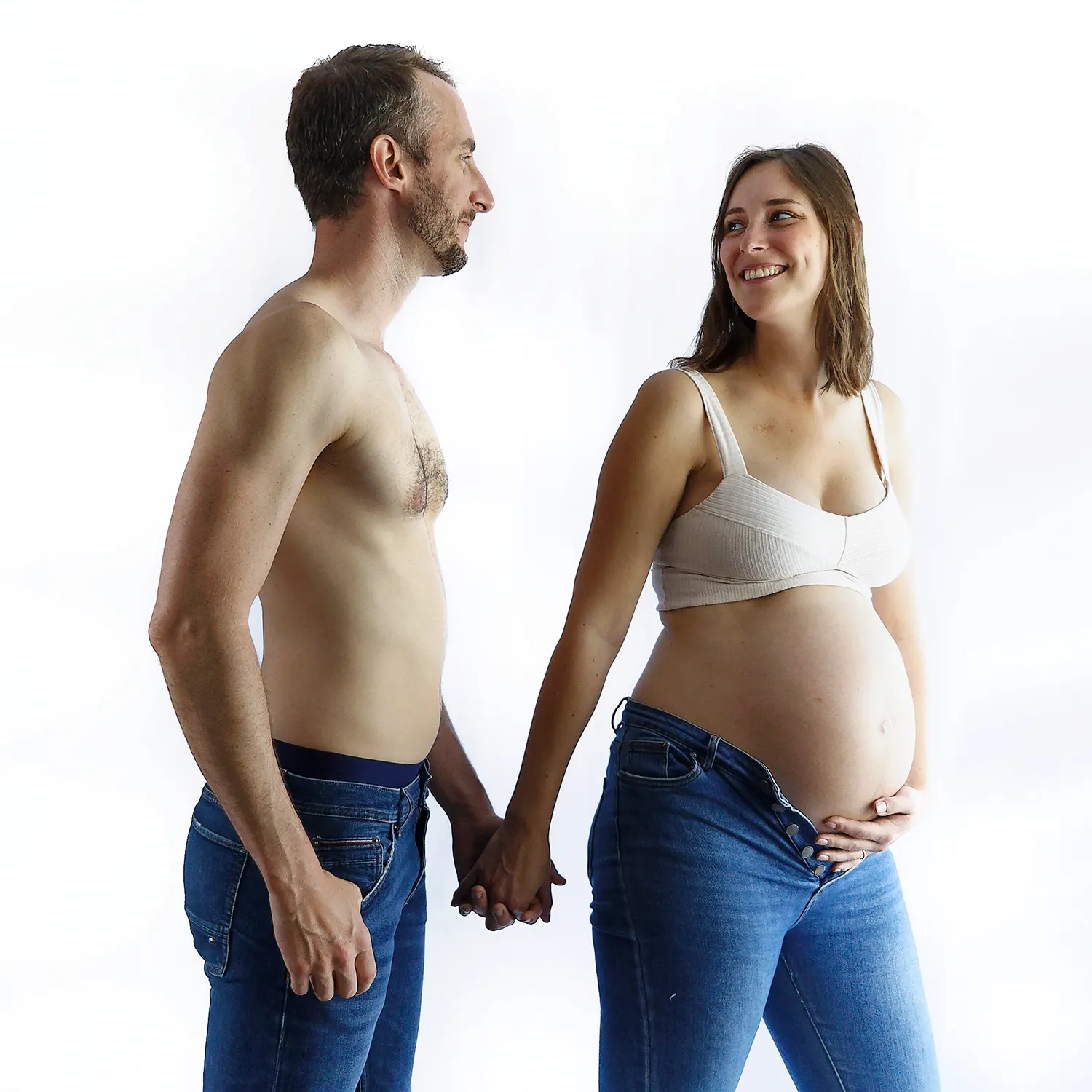 Séance photo de grossesse en studio lumineux. Un couple se tenant la main et se regardant avec tendresse, sur un fond blanc.