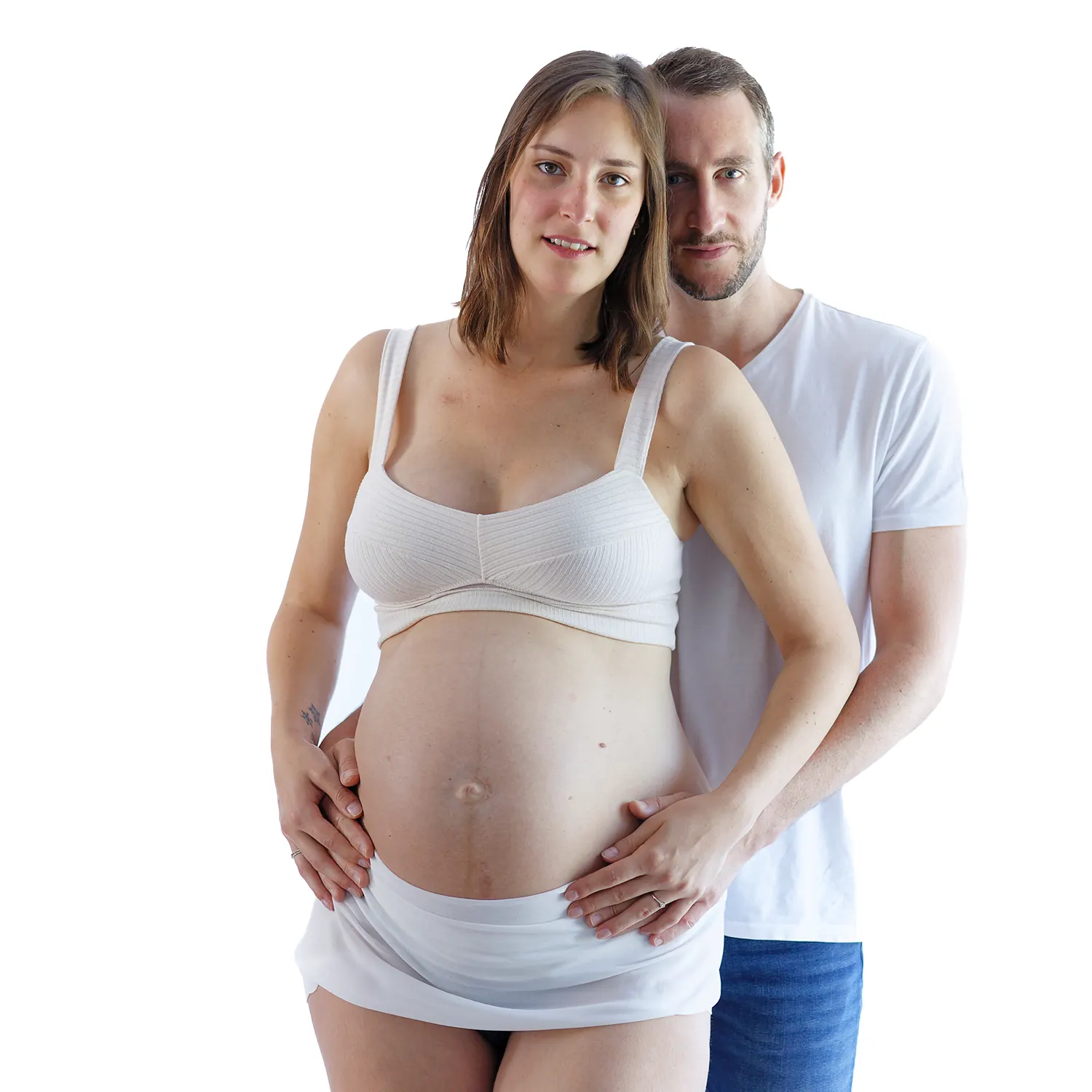 : Portrait de grossesse en studio lumineux d'un couple. Le futur papa enlaçant la future maman par-derrière, les mains posées sur son ventre, sur un fond blanc.