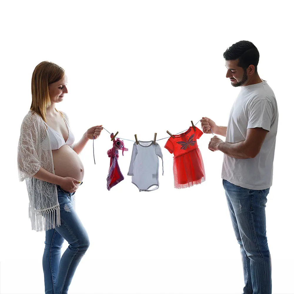 Séance photo de grossesse en studio lumineux. Un couple souriant tient une corde à linge avec de petits vêtements de bébé, symbolisant l'arrivée prochaine.