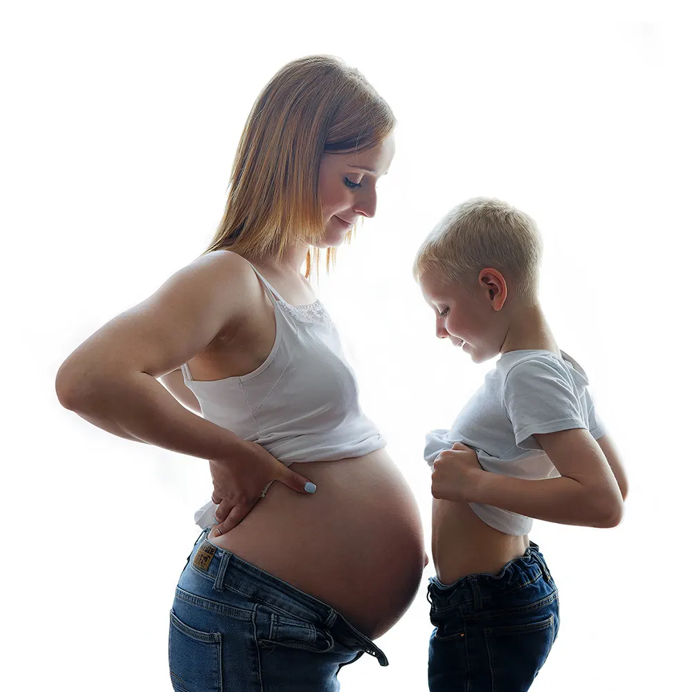 Séance photo de grossesse en studio lumineux. Une future maman et son jeune fils regardent leurs ventres avec tendresse, sur un fond blanc.