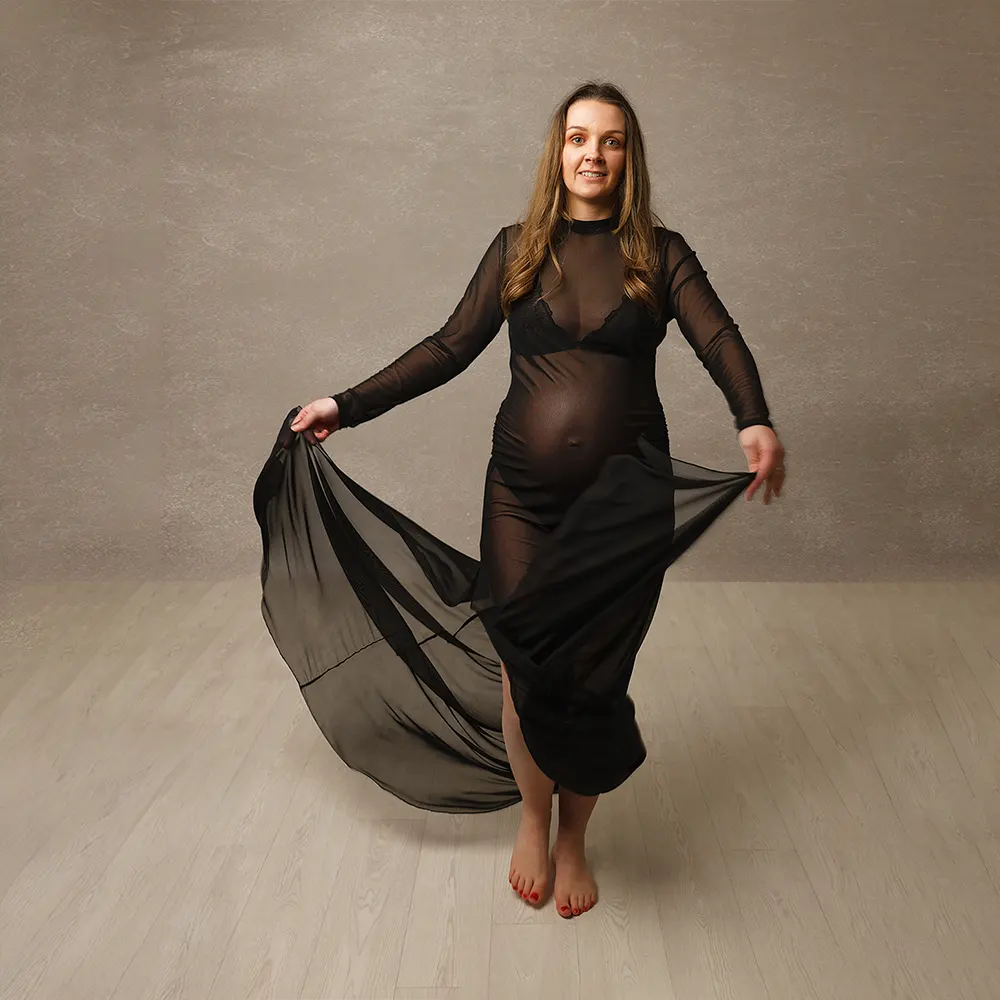 Séance photo de maternité en studio. La future maman porte une robe longue noire transparente pour mettre en évidence ses rondeurs. Son mouvement est figé mais rend la photo dynamique.