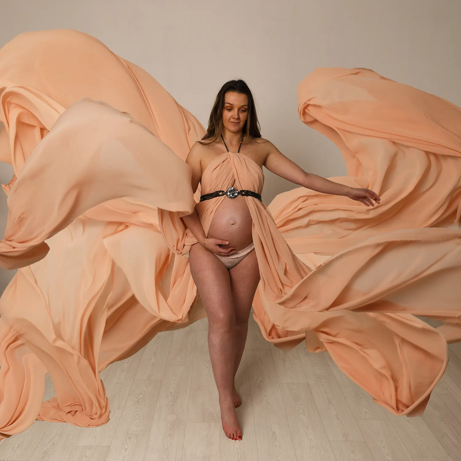 Portrait de grossesse en studio. Une future maman porte une robe fluide couleur pêche, le tissu flottant autour d'elle, tenant son ventre rond.