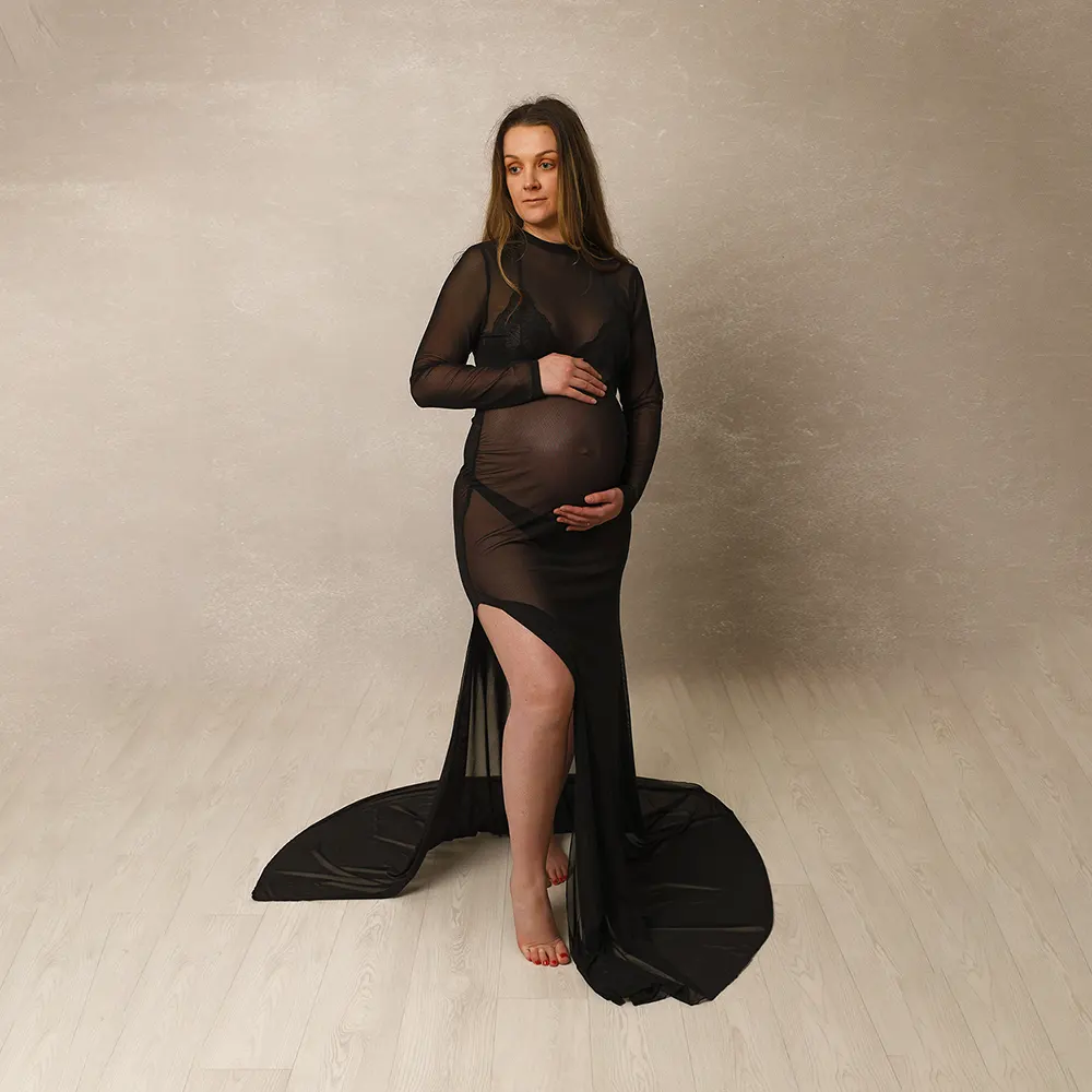 Portrait de grossesse en studio d'une future maman. Elle porte une élégante robe longue noire transparente et pose en tenant son ventre.
