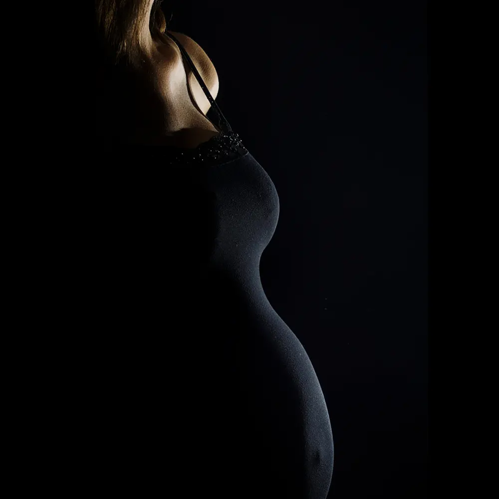 Portrait de grossesse en studio. La silhouette élégante d'une future maman est éclairée pour mettre en valeur le profil de son ventre rond, sur un fond noir.