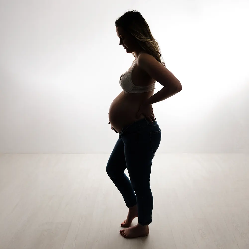 Séance photo de grossesse en studio. La silhouette d'une femme enceinte est mise en valeur par un éclairage de fond, soulignant son profil et son ventre rond.