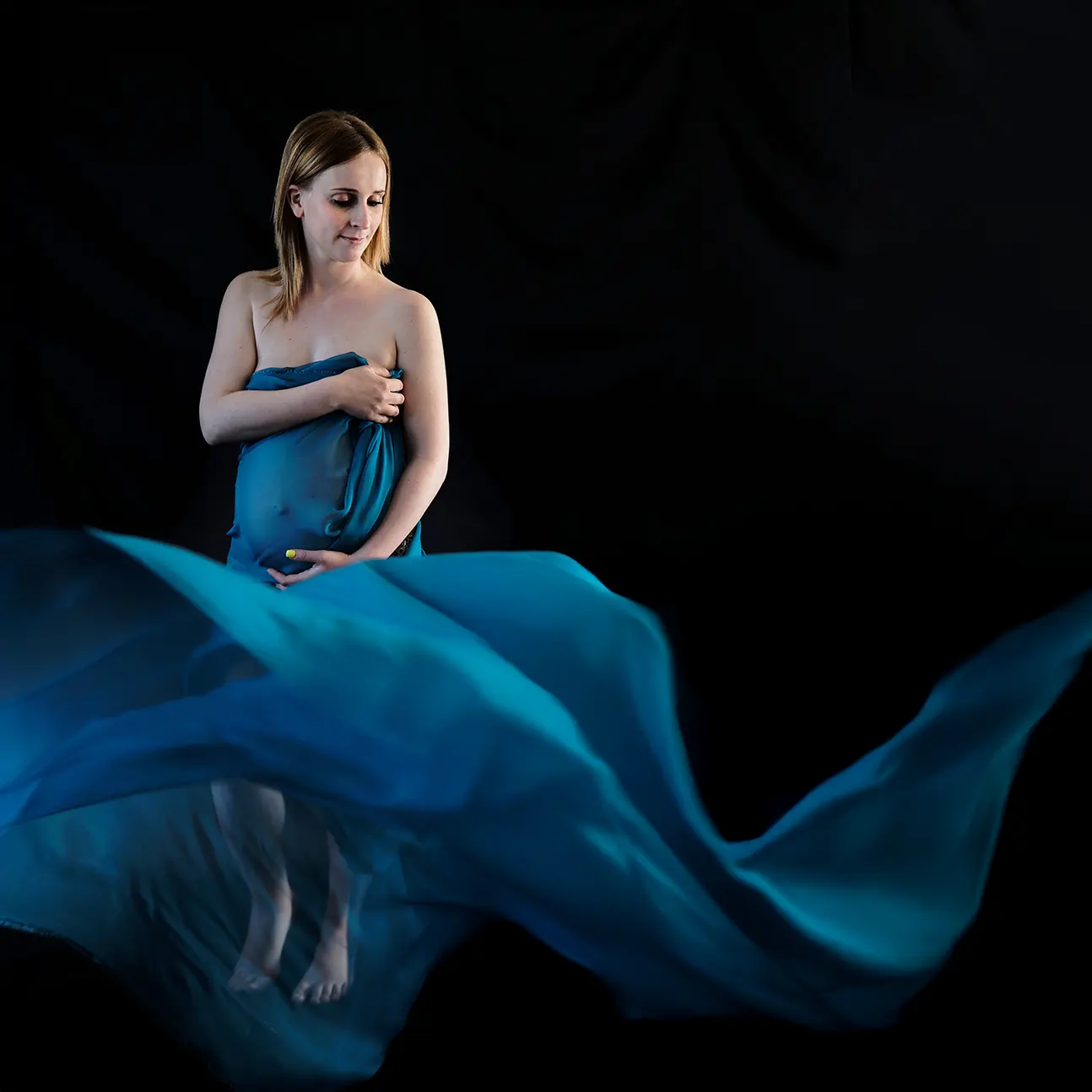 Séance photo de maternité en studio. Une future maman drapée dans un voilage bleu volant, tenant son ventre avec un fond noir