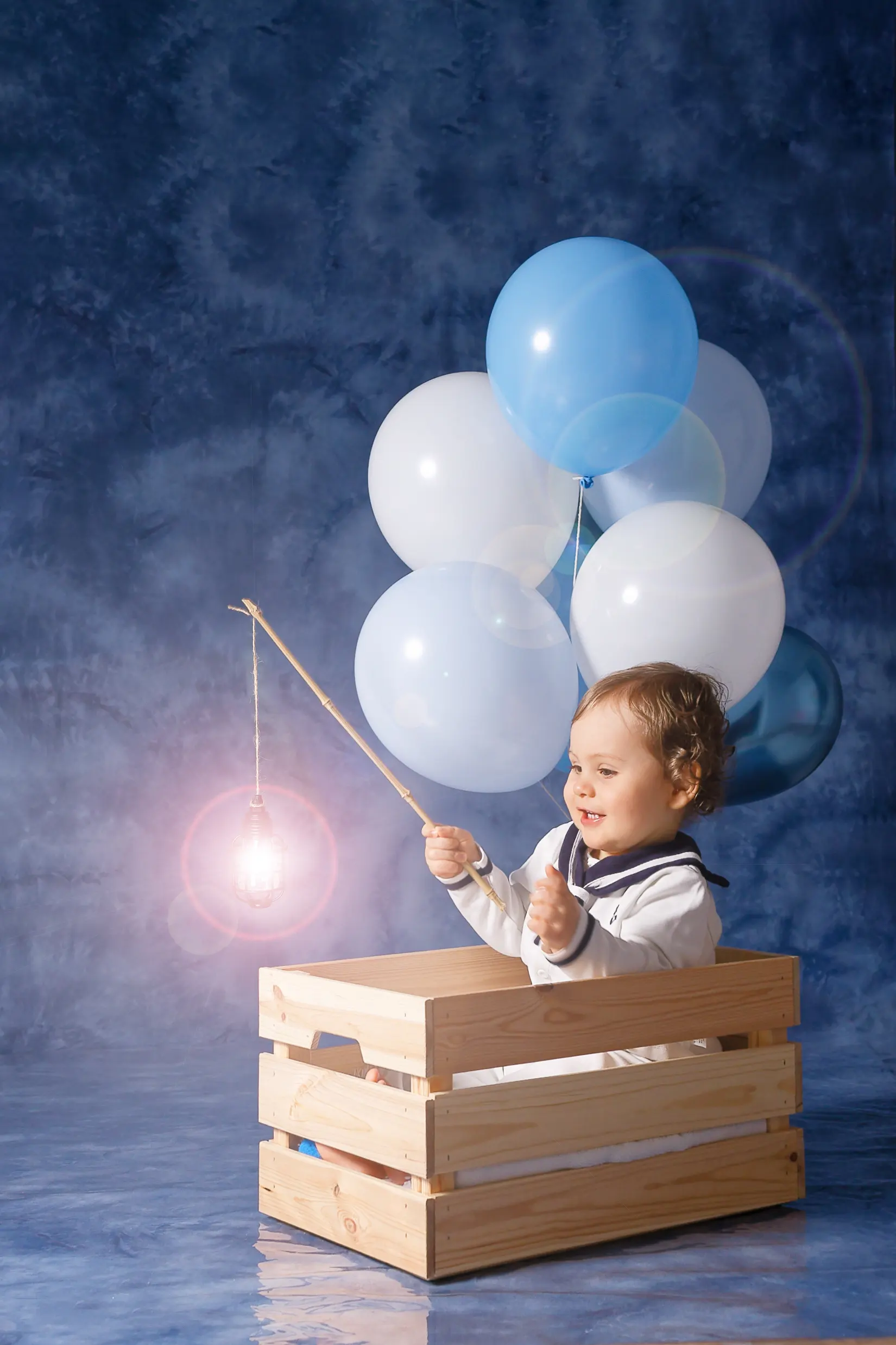 Un bébé souriant assis dans une caisse en bois, vêtu d'un costume de marin. Il tient une canne à pêche avec une ampoule lumineuse au bout. Des ballons bleus et blancs flottent derrière lui.