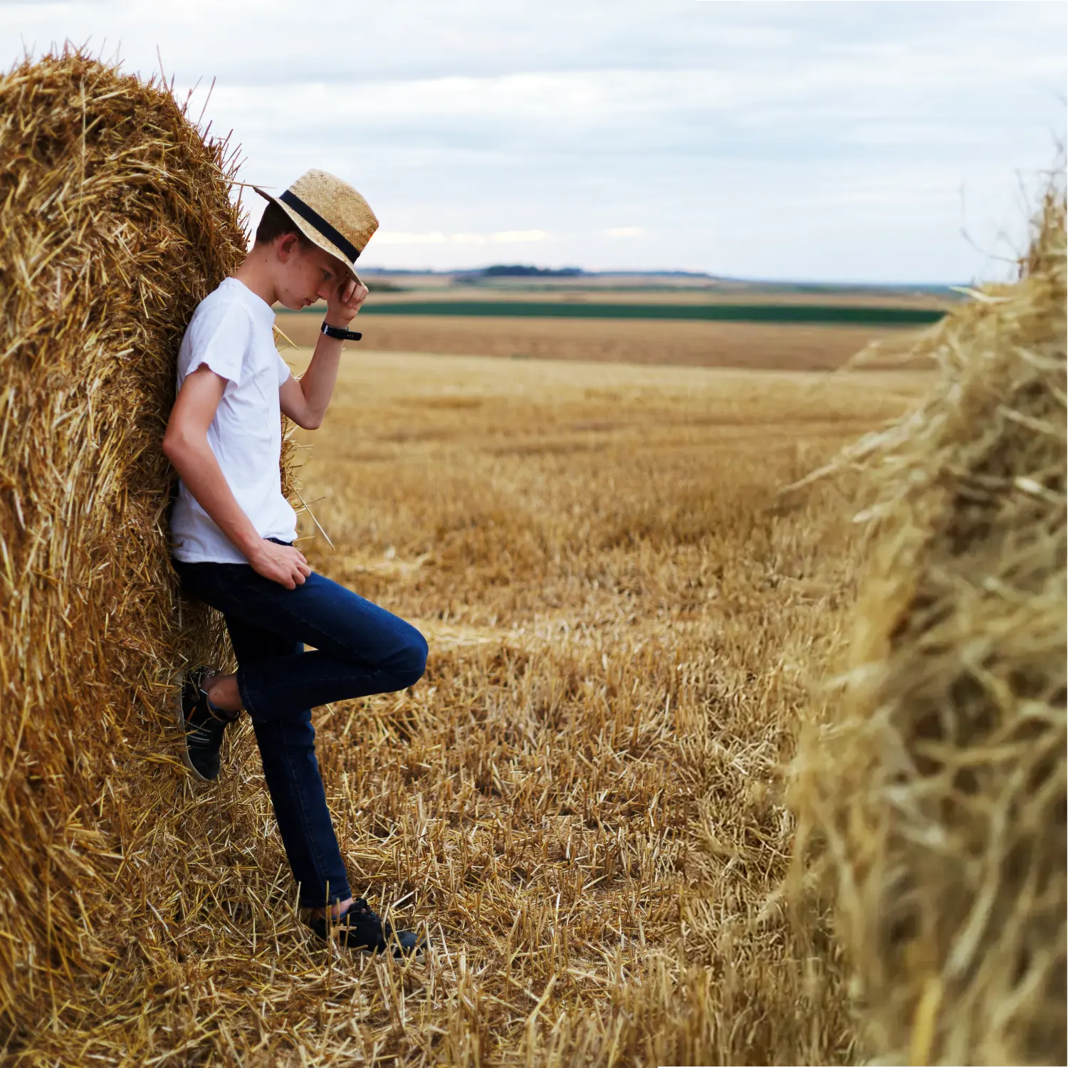 Portrait d'un adolescent avec un chapeau, posant avec une expression pensive contre une botte de foin.