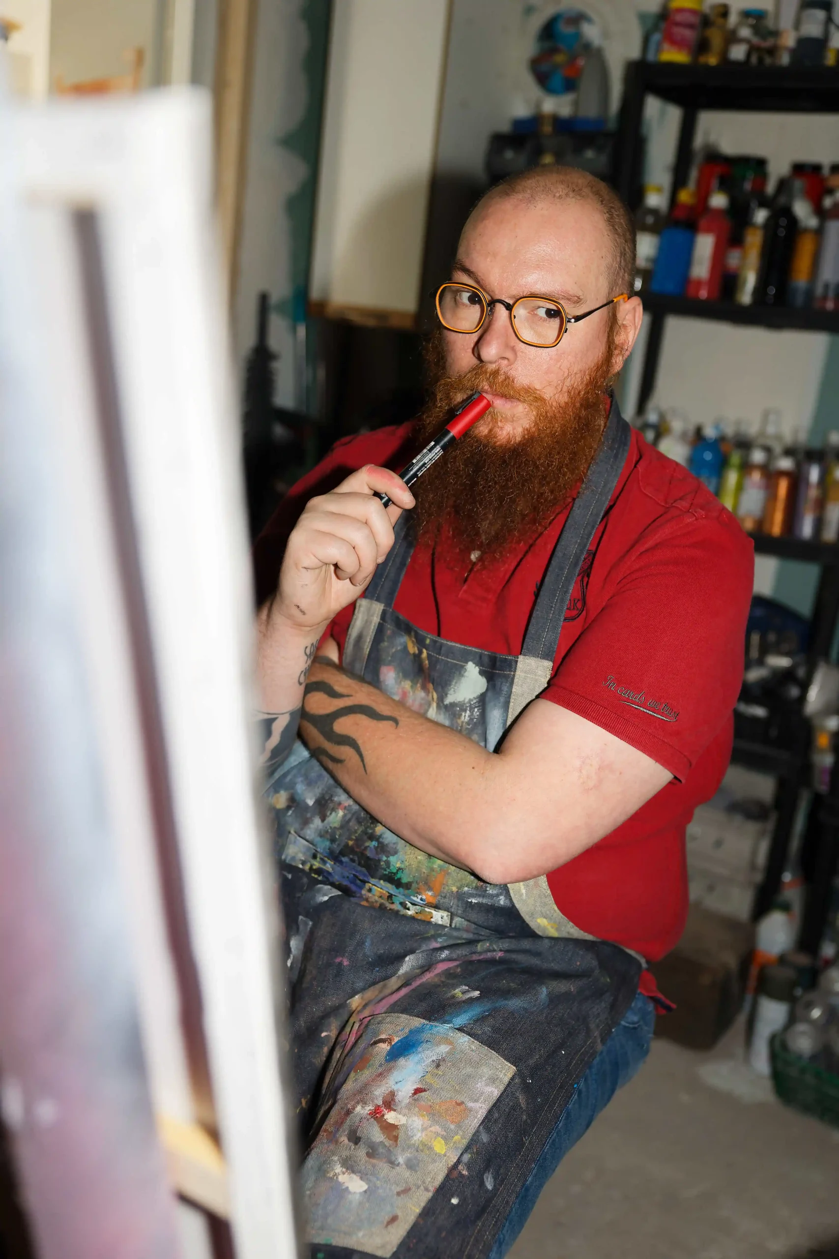 Un artiste-peintre assis dans son atelier, tenant un stylo-feutre près de sa bouche, l'air pensif devant un chevalet.