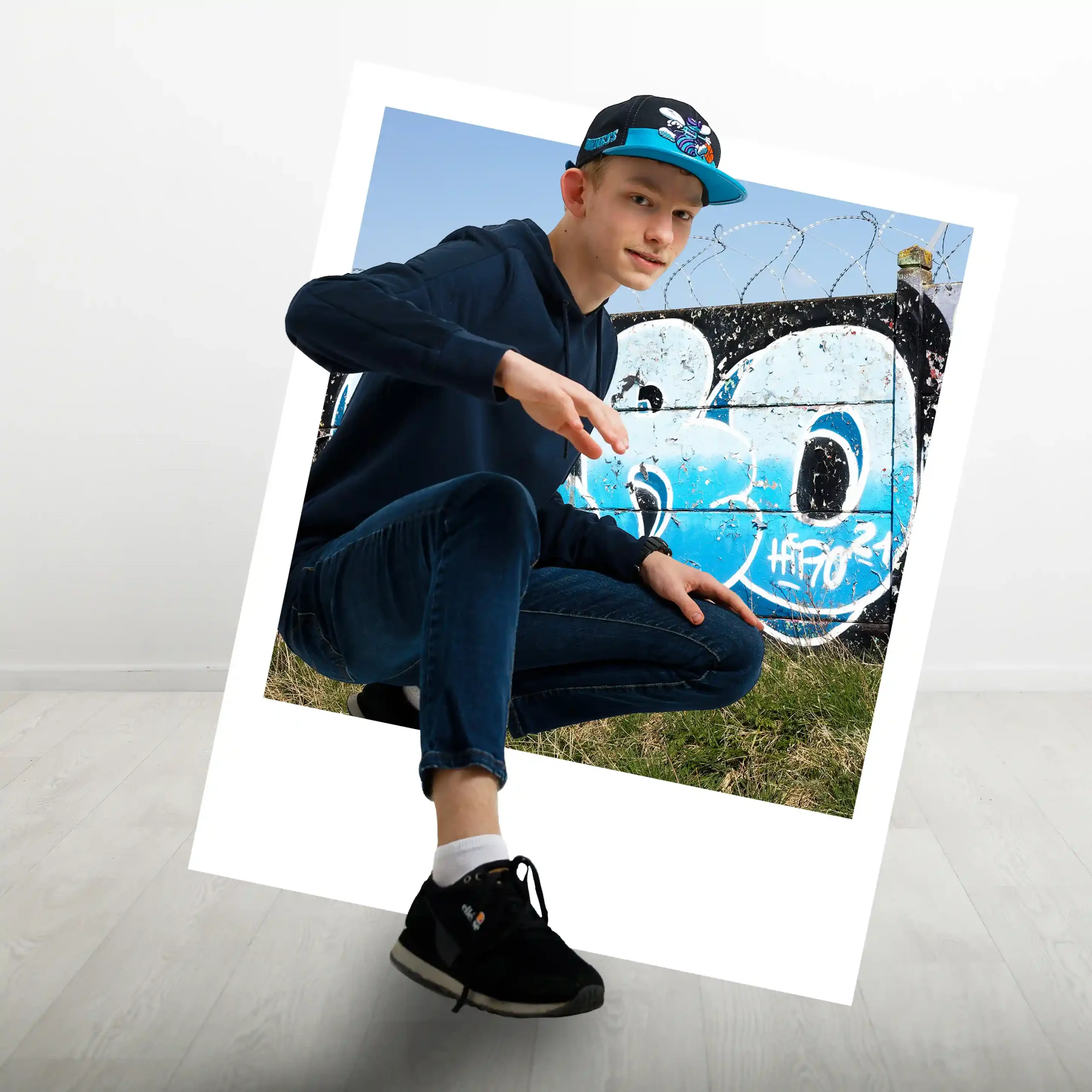 Photomontage d'un adolescent accroupi devant un mur tagué. Il porte une casquette et un sweat-shirt bleu, avec ses genoux et ses pieds en dehors du cadre photo de type Polaroid.