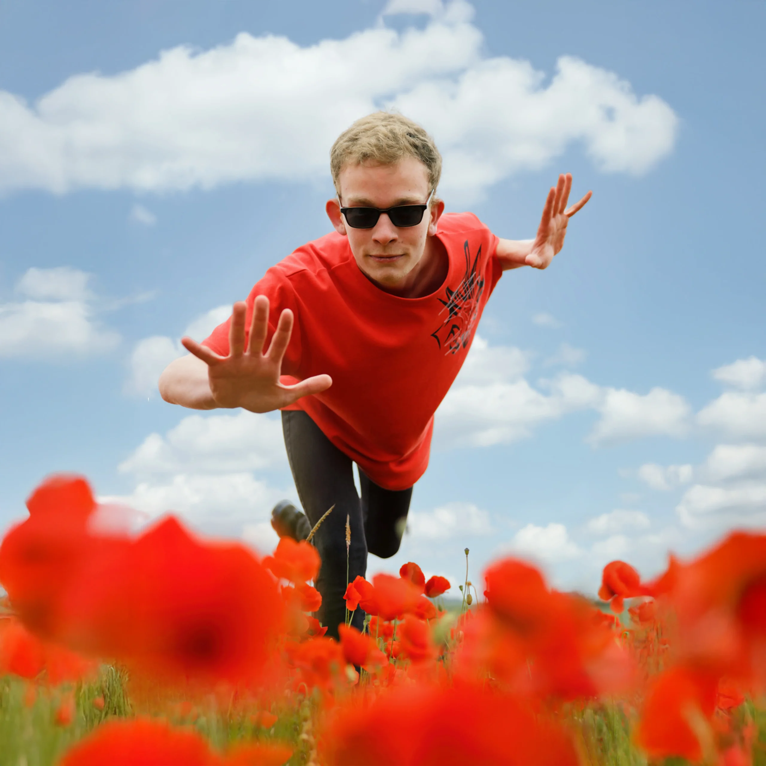 Portrait dynamique d'un homme en t-shirt rouge et lunettes de soleil, sautant dans un champ de coquelicots sous un ciel bleu.